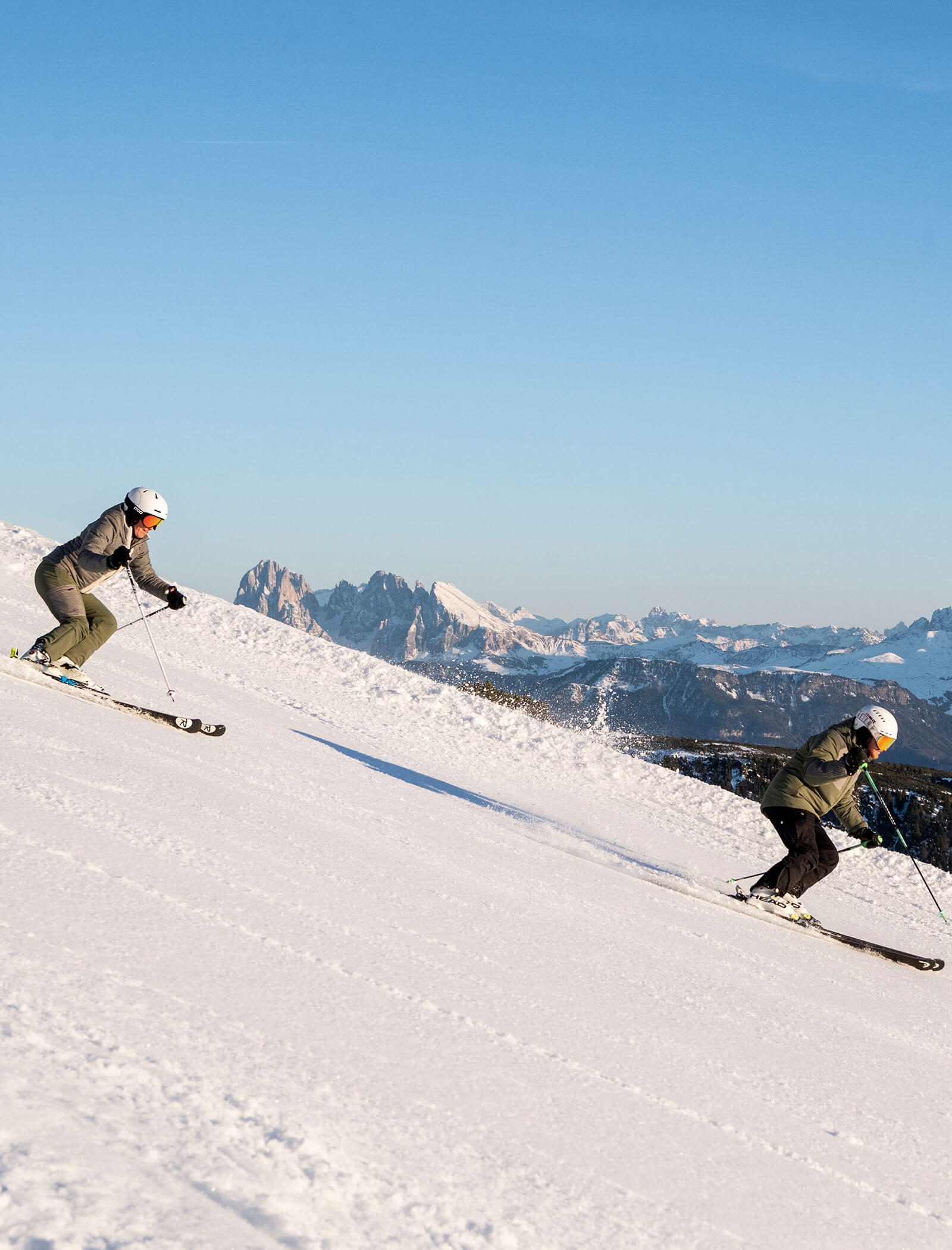 Due persone che scendono con gli sci lungo un pendio innevato, con le montagne e il cielo azzurro sullo sfondo. - Hotel Kristiania