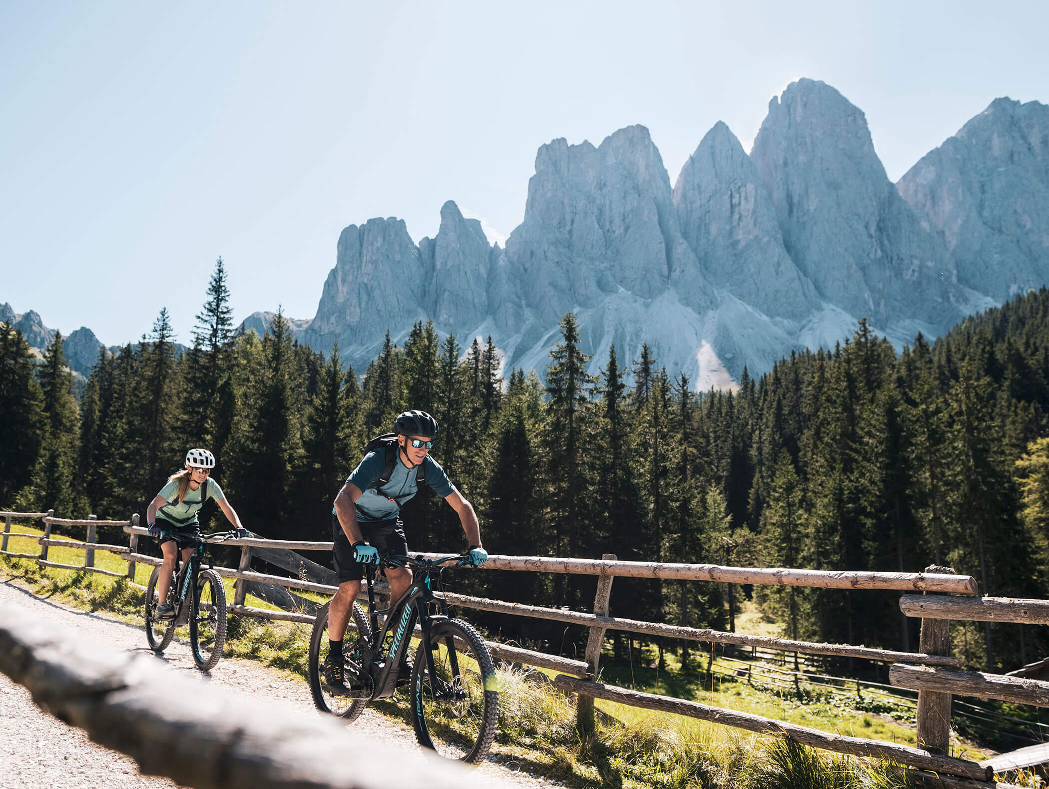 Two cyclists ride mountain bikes on a dirt trail with pine trees and rocky mountains in the background. - Hotel Kristiania