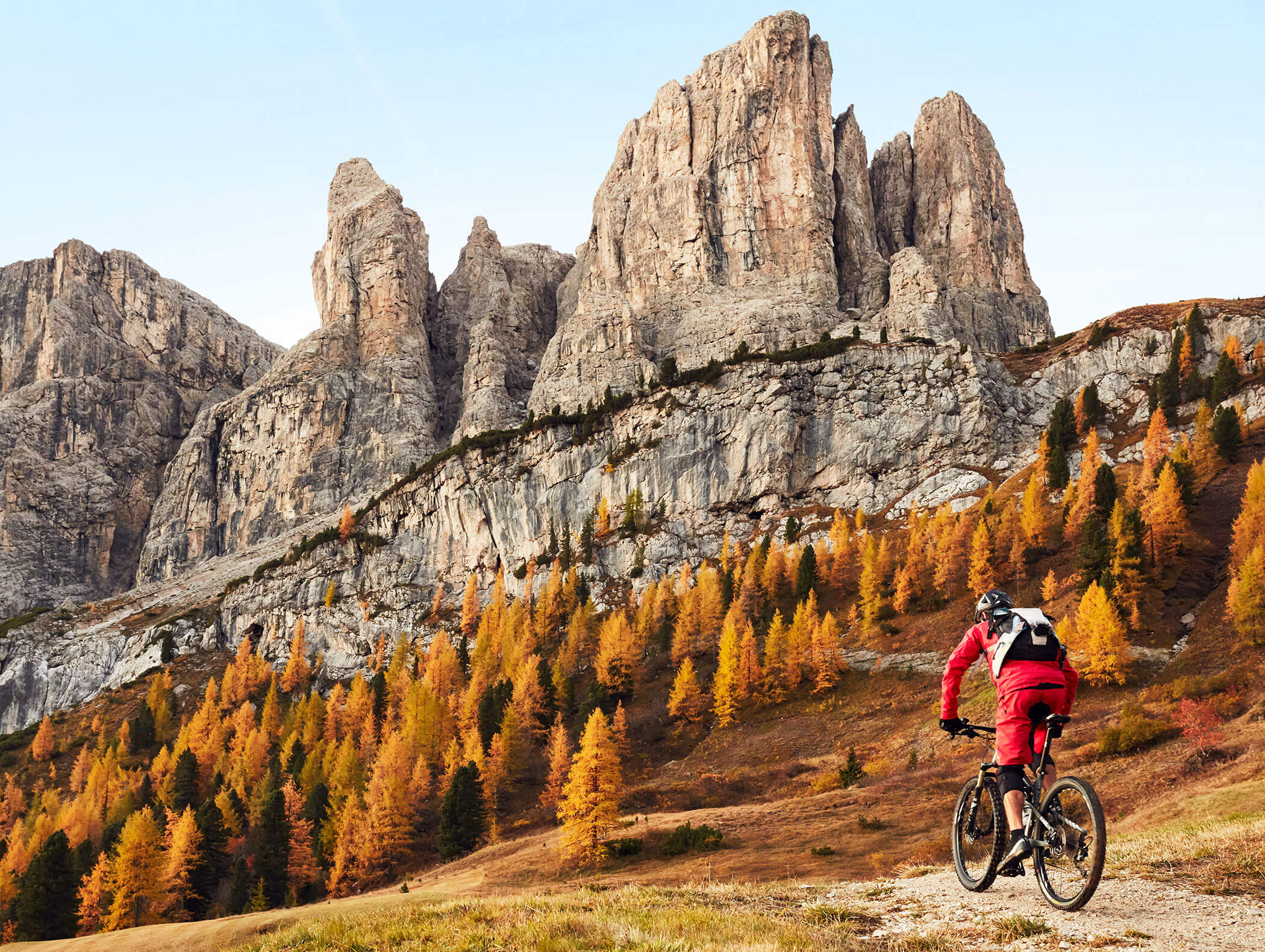 A cyclist in red rides toward tall, rocky mountains with autumn trees in the foreground. - Hotel Kristiania