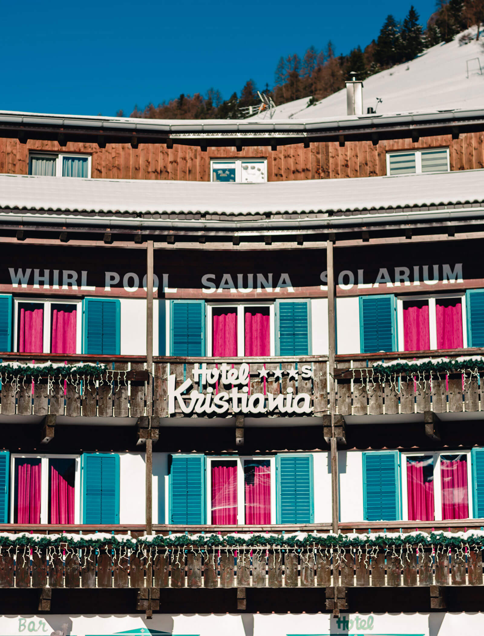 Hotel with wooden balconies, pink curtains, and a sign reading Hotel Krushania in a snowy mountain setting. - Hotel Kristiania