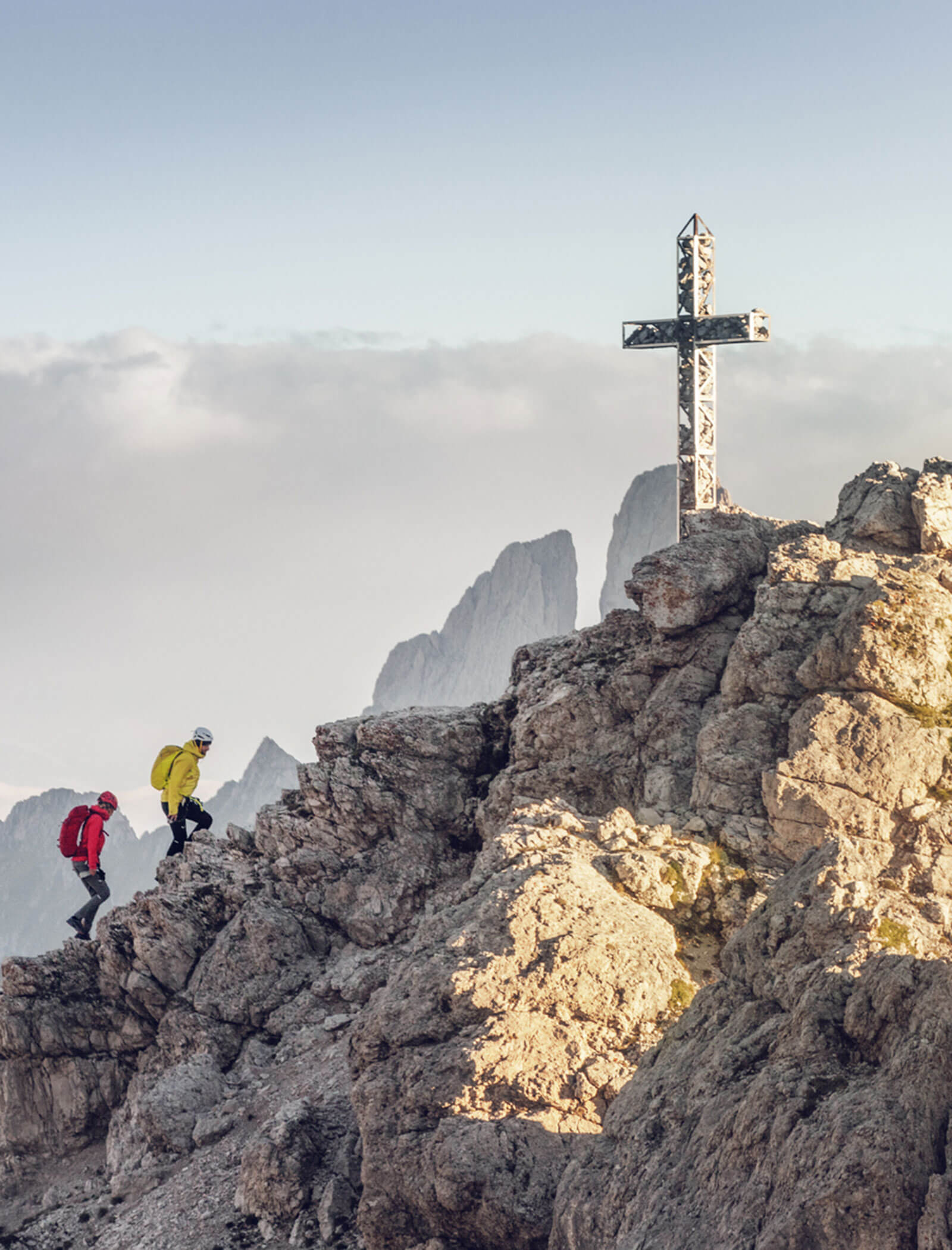 Due escursionisti si arrampicano su un terreno roccioso verso una grande croce in cima a una montagna, con le vette e le nuvole sullo sfondo. - Hotel Kristiania