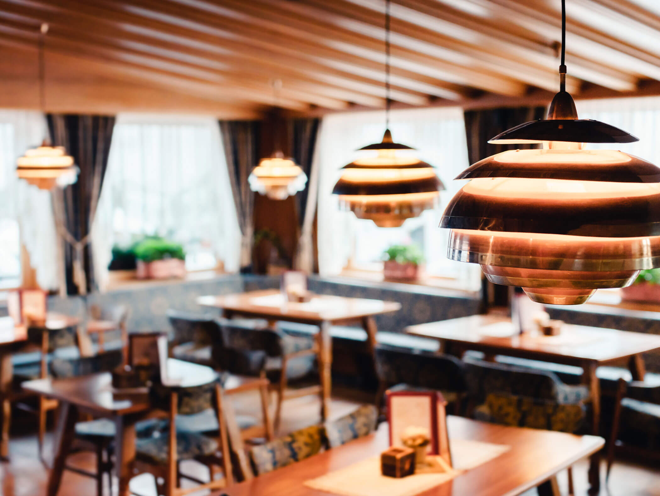Modern pendant lights hanging above empty wooden tables in a cozy, sunlit restaurant with large windows. - Hotel Kristiania