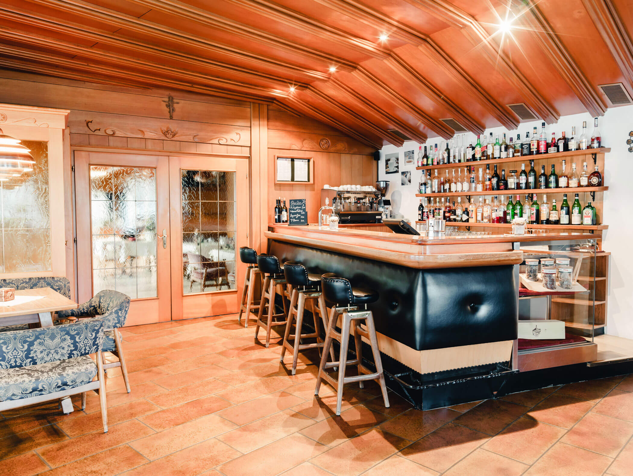 Cozy bar with a wooden ceiling, stocked shelves, cushioned stools, and tables with patterned seating. - Hotel Kristiania