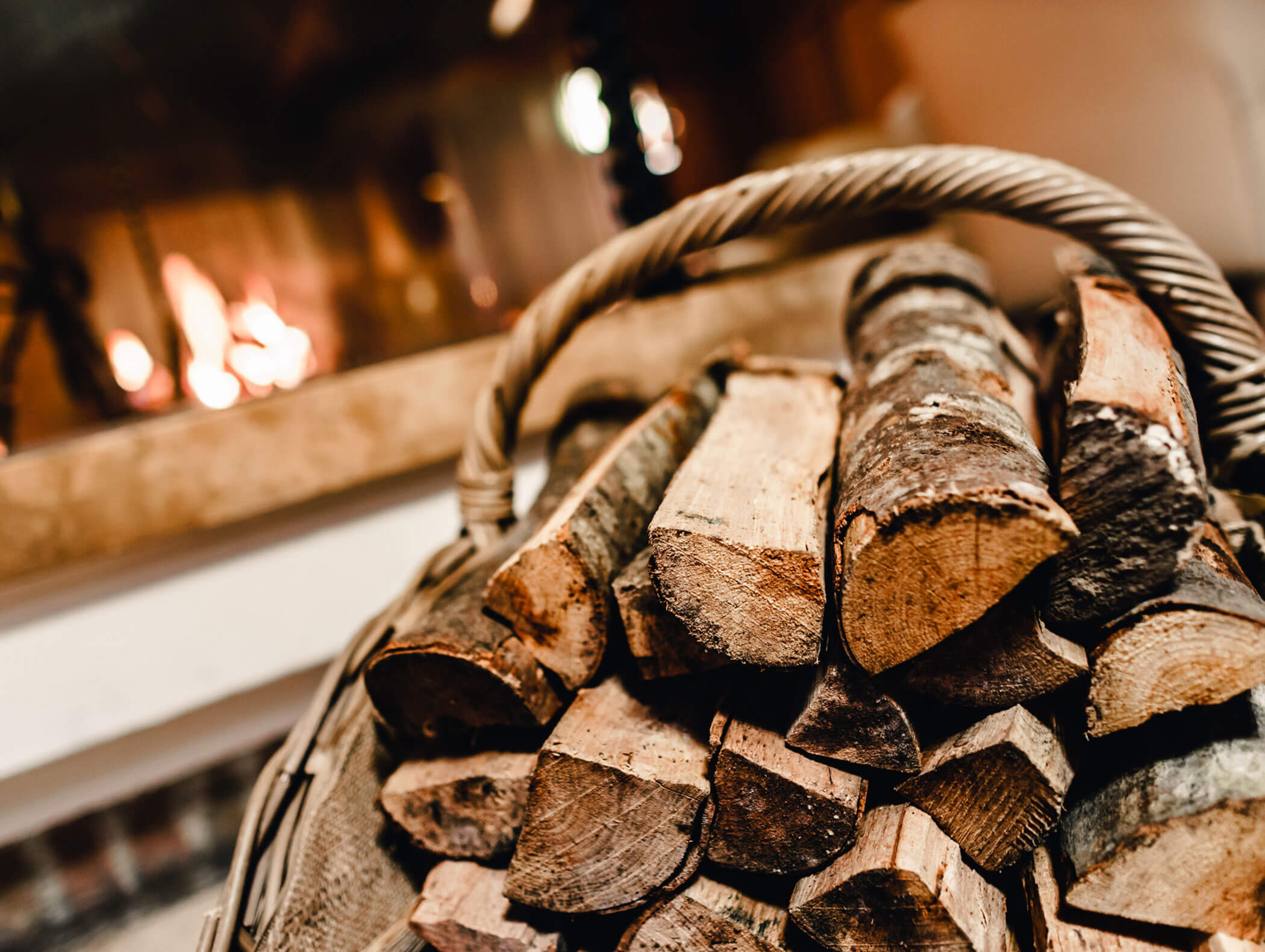 A basket of firewood sits near a fireplace with a fire burning in the background. - Hotel Kristiania