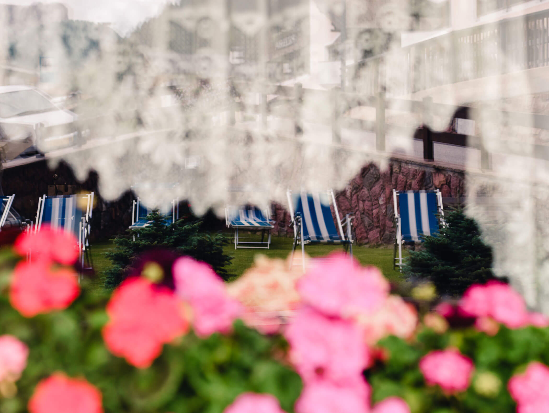 Blurred flowers and striped deck chairs seen through a lace-covered window, creating a dreamy effect. - Hotel Kristiania