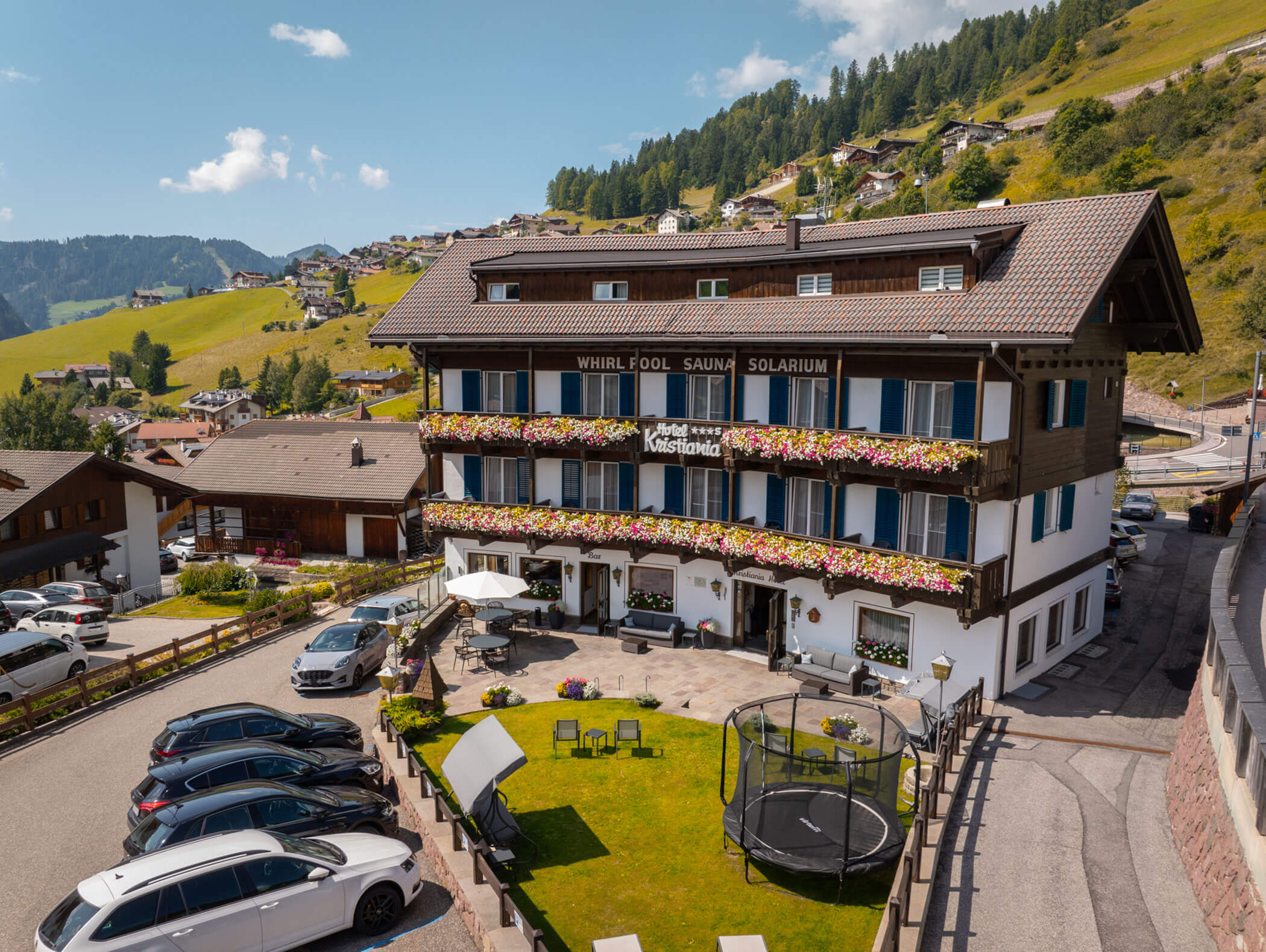 Chalet-style hotel with flower balconies, parked cars, and mountain village scenery in the background. - Hotel Kristiania
