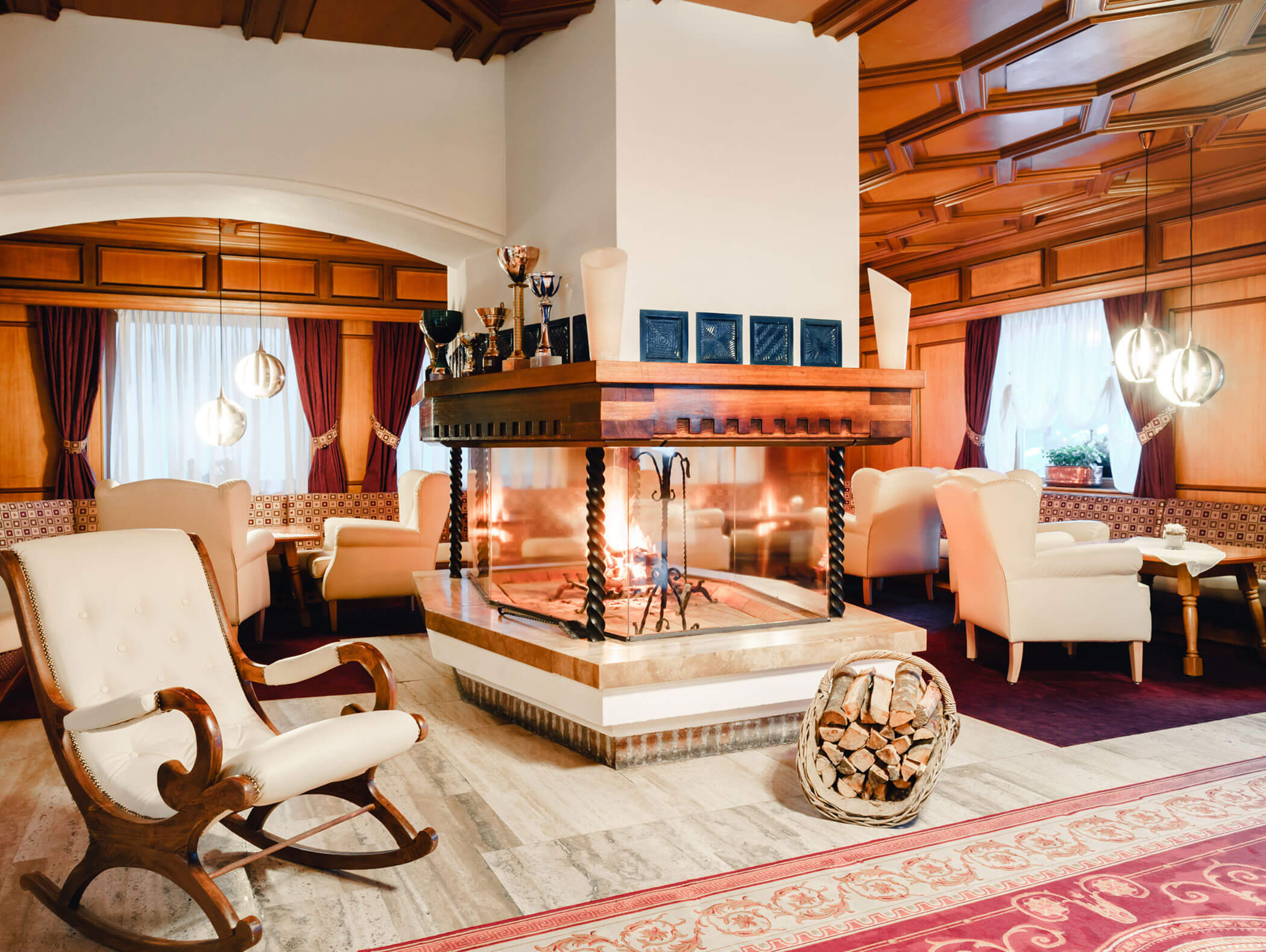 Cozy lounge with a central fireplace, armchairs, a rocking chair, and stacked firewood in a basket. - Hotel Kristiania