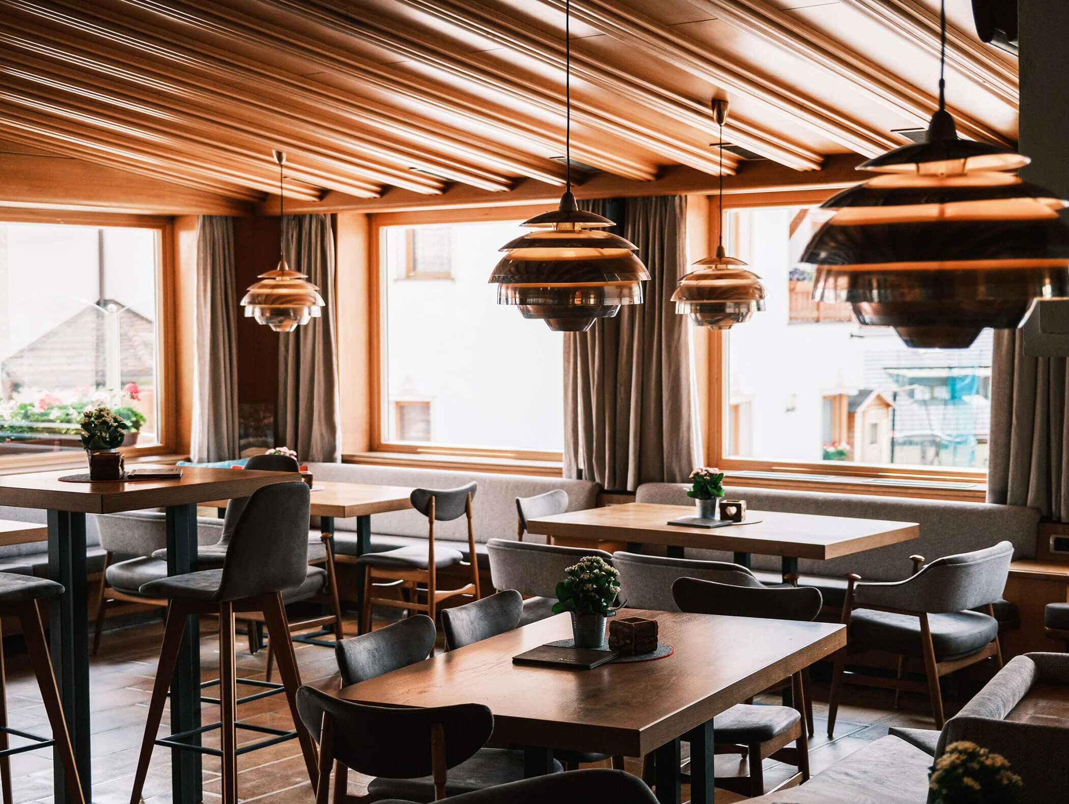A cozy, modern restaurant with wooden tables, chairs, pendant lights, and large windows letting in natural light. - Hotel Kristiania