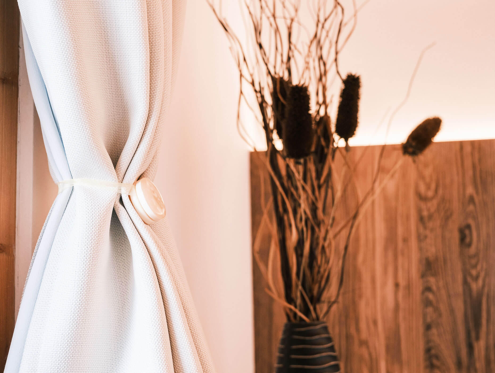 White curtain tied back near wooden panel wall; vase with dried branches in the background. - Hotel Kristiania