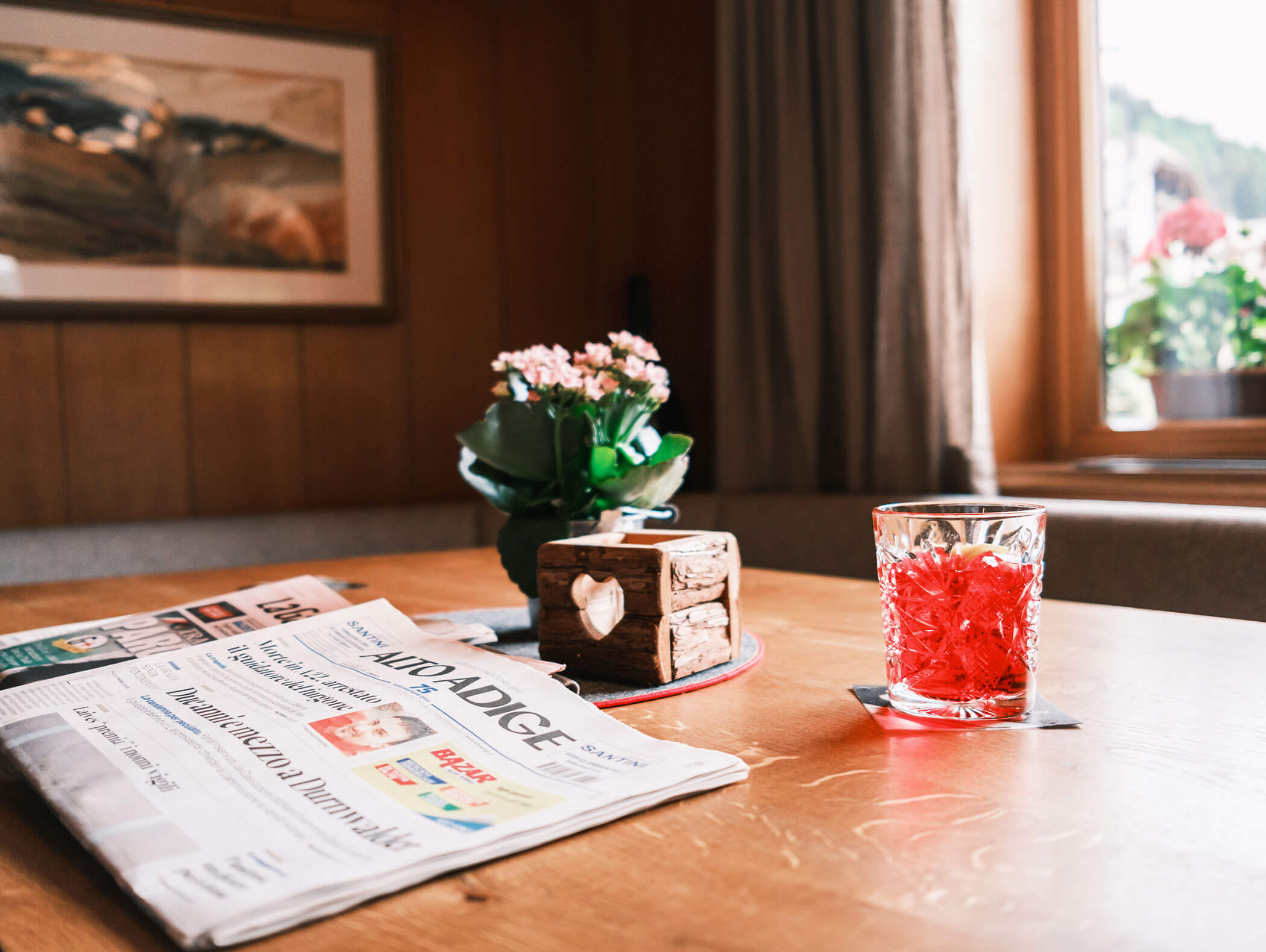 A glass of red drink, a newspaper, and a small potted plant on a wooden table by a window. - Hotel Kristiania