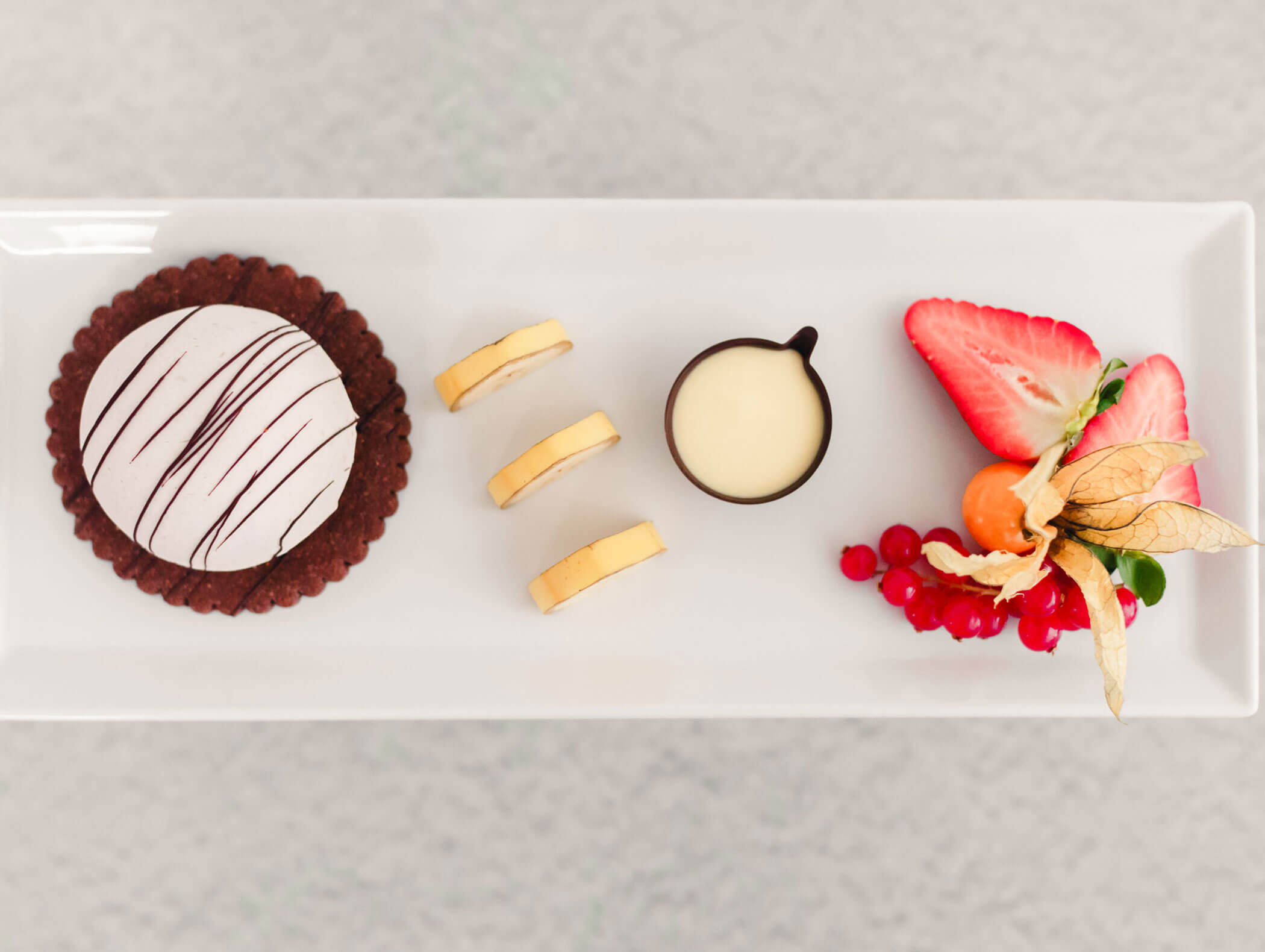 A chocolate tart, banana slices, cream, and fresh fruit arranged on a rectangular white plate. - Hotel Kristiania