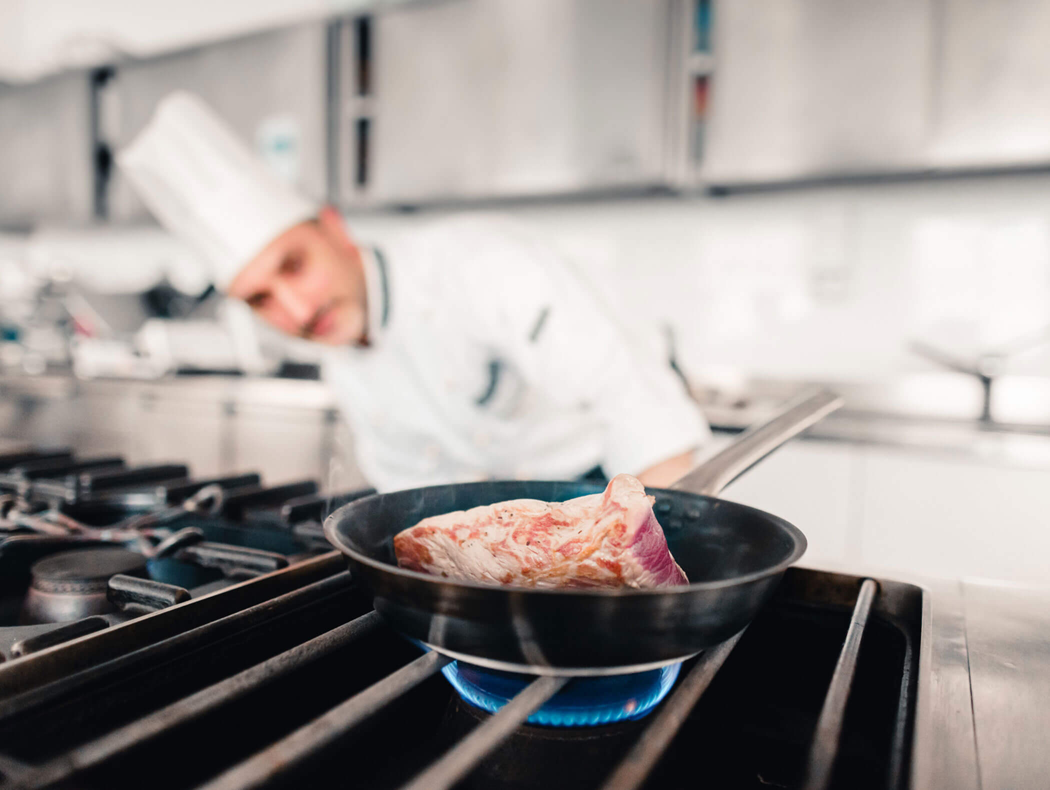 A chef in a kitchen cooks a raw steak in a frying pan over a gas stove flame. - Hotel Kristiania