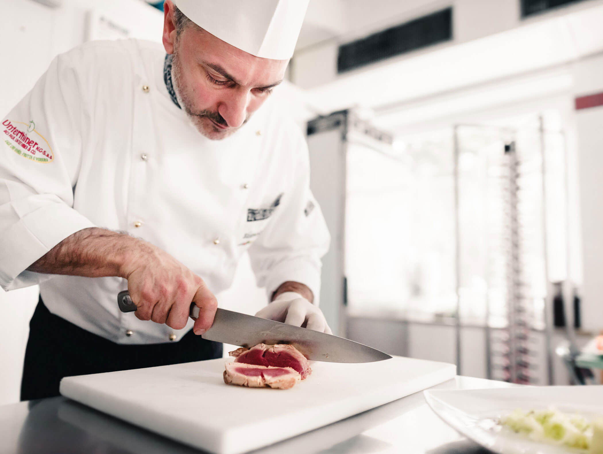 Chef in a white uniform slicing raw meat on a cutting board in a professional kitchen. - Hotel Kristiania