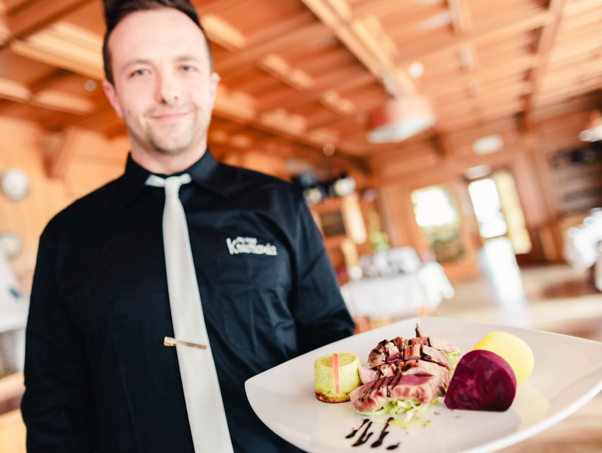 A waiter in a black shirt serves a gourmet dish on a white plate in a warmly lit restaurant. - Hotel Kristiania
