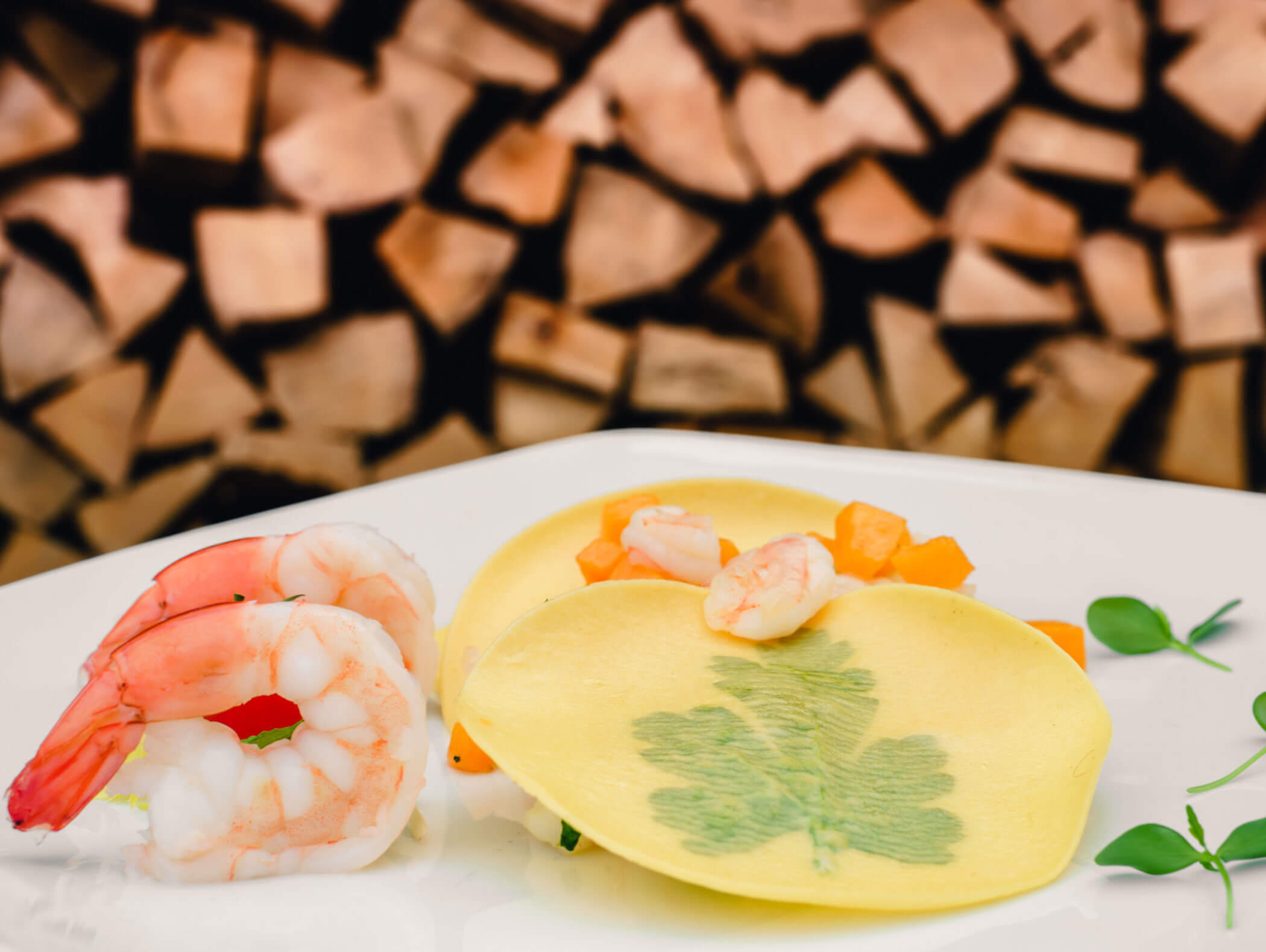 Two shrimp beside ravioli garnished with herbs, served on a white plate with stacked wood in the background. - Hotel Kristiania