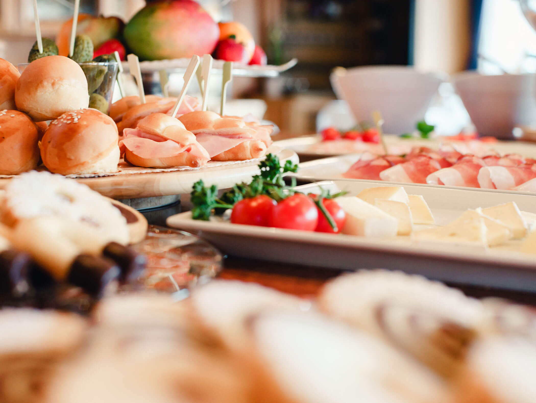 Assorted sandwiches, cheeses, and fresh vegetables arranged on a buffet table with a fruit platter in the background. - Hotel Kristiania