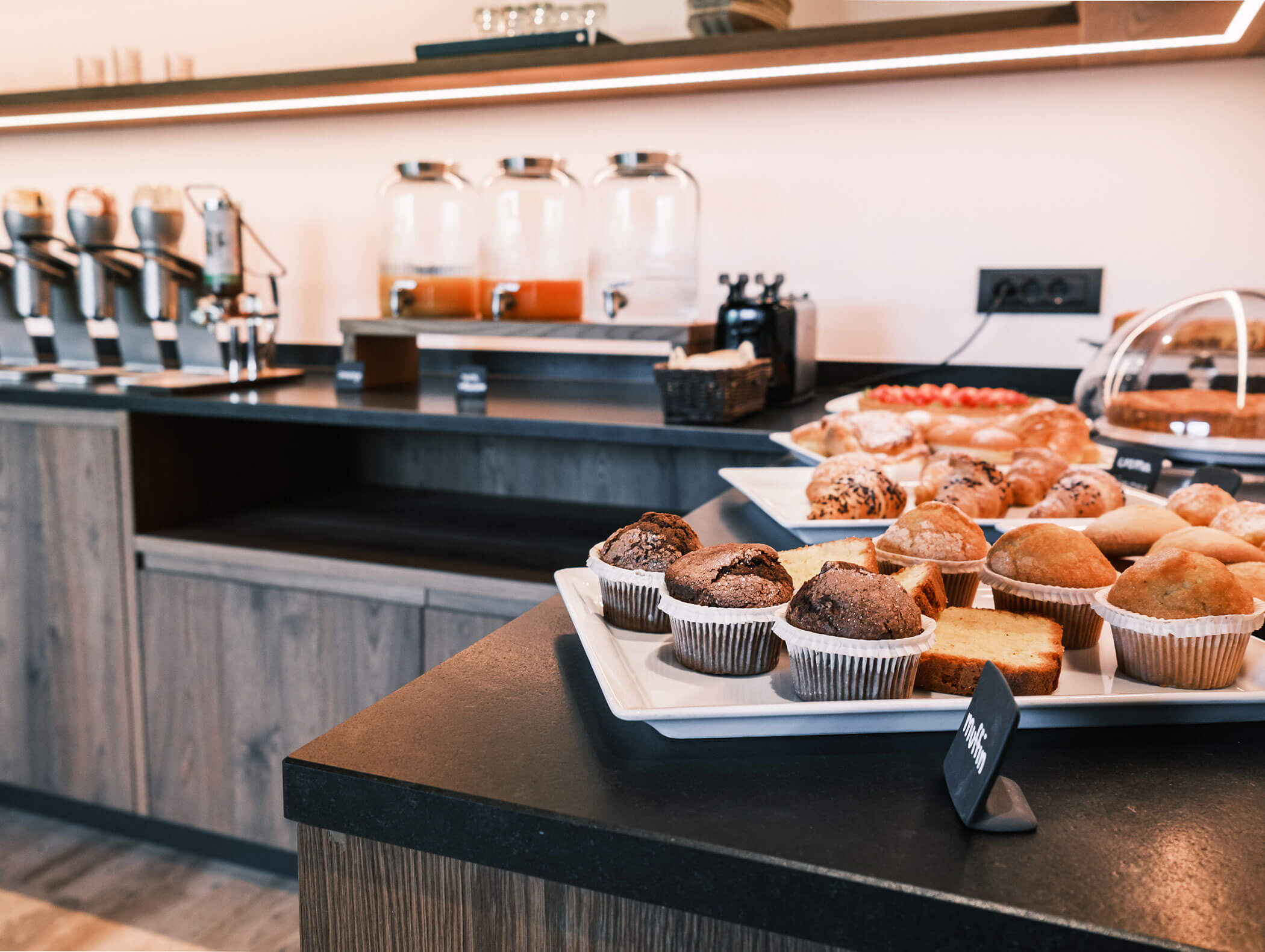 Assorted muffins and pastries on trays in a modern breakfast buffet with juice dispensers in the background. - Hotel Kristiania