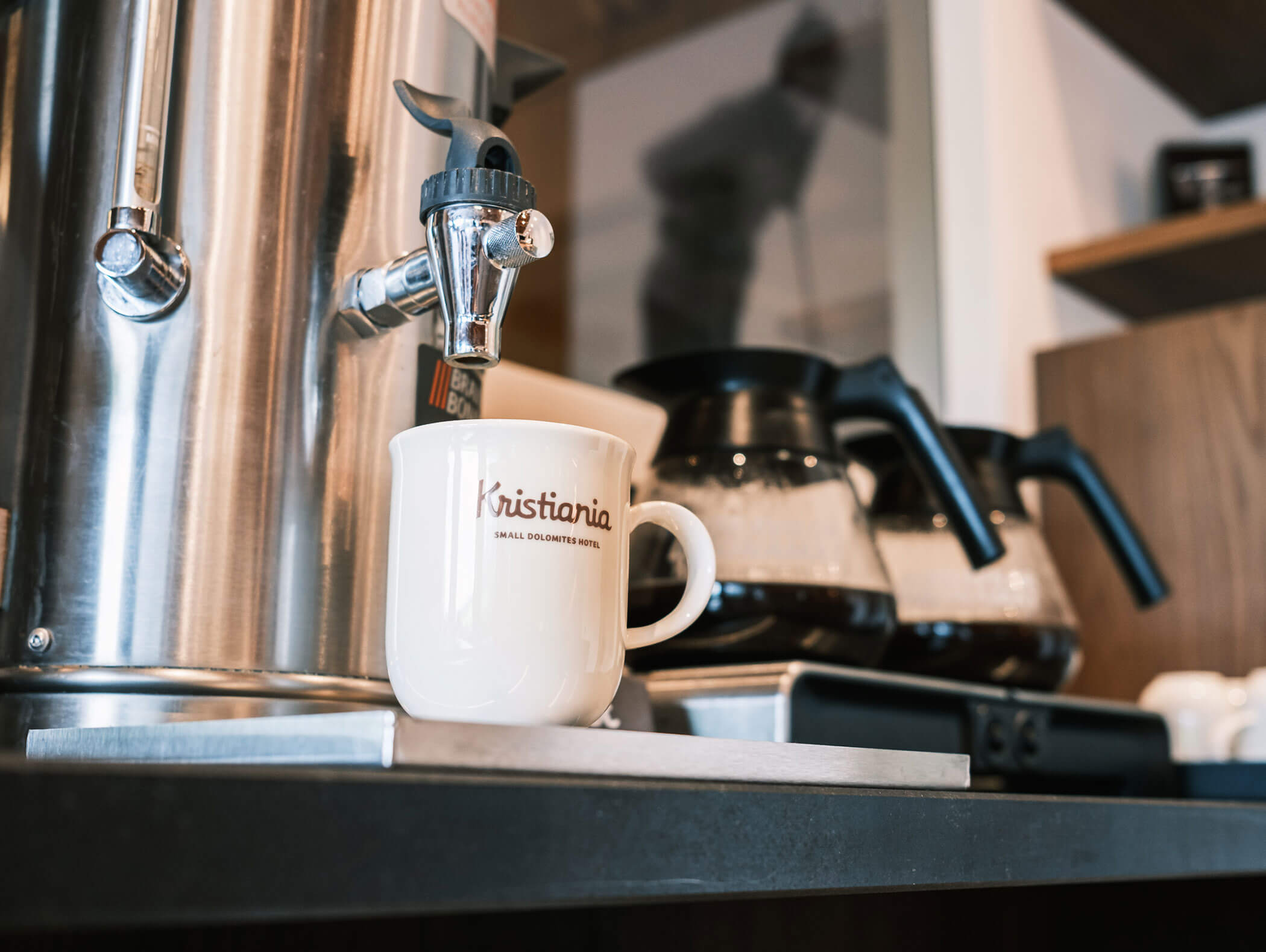 White coffee mug by a coffee urn, with two glass coffee pots and wooden shelves in the background. - Hotel Kristiania