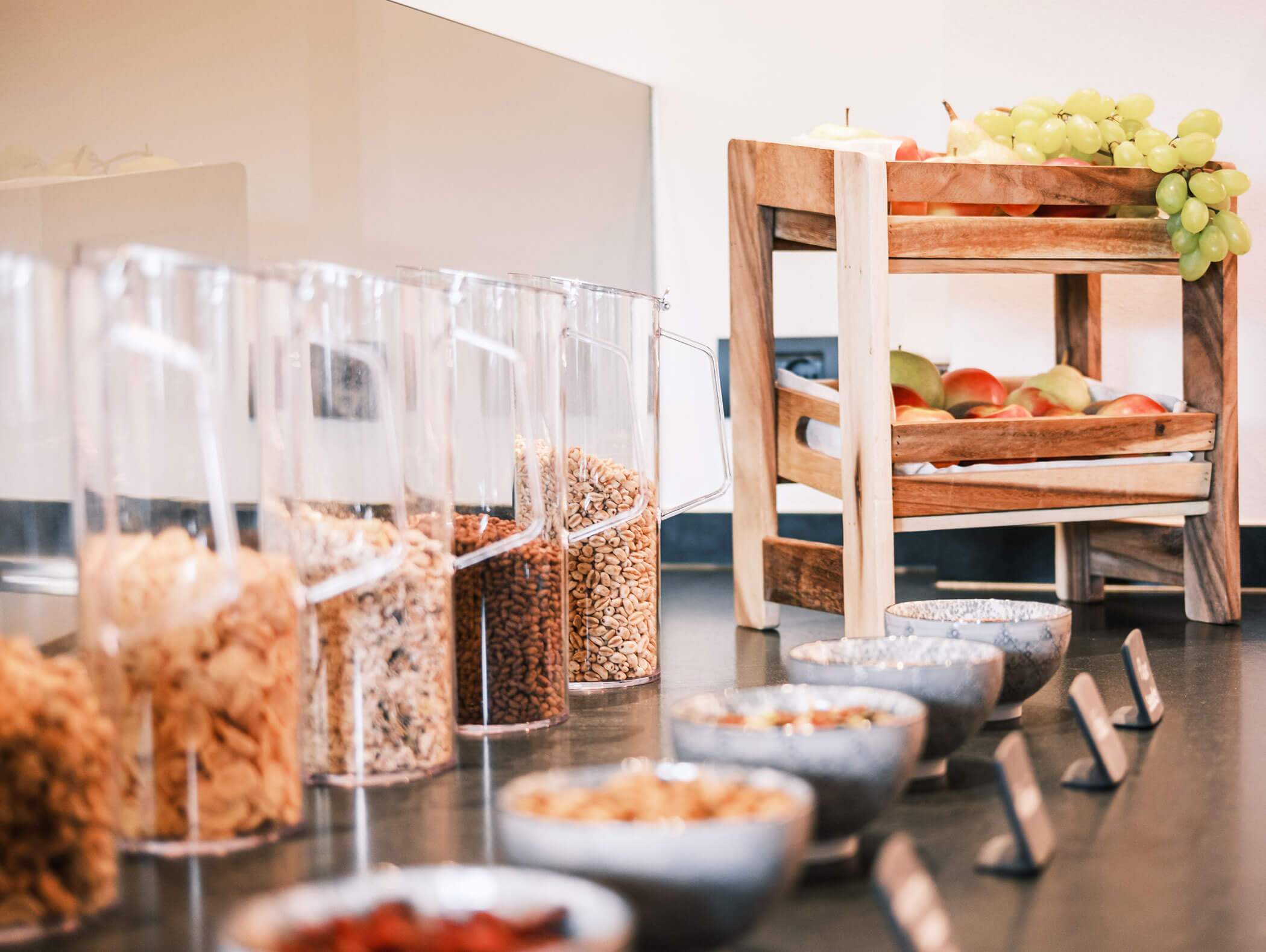 Clear food dispensers with cereal and nuts beside bowls and a wooden stand holding fresh fruit, including grapes and apples. - Hotel Kristiania