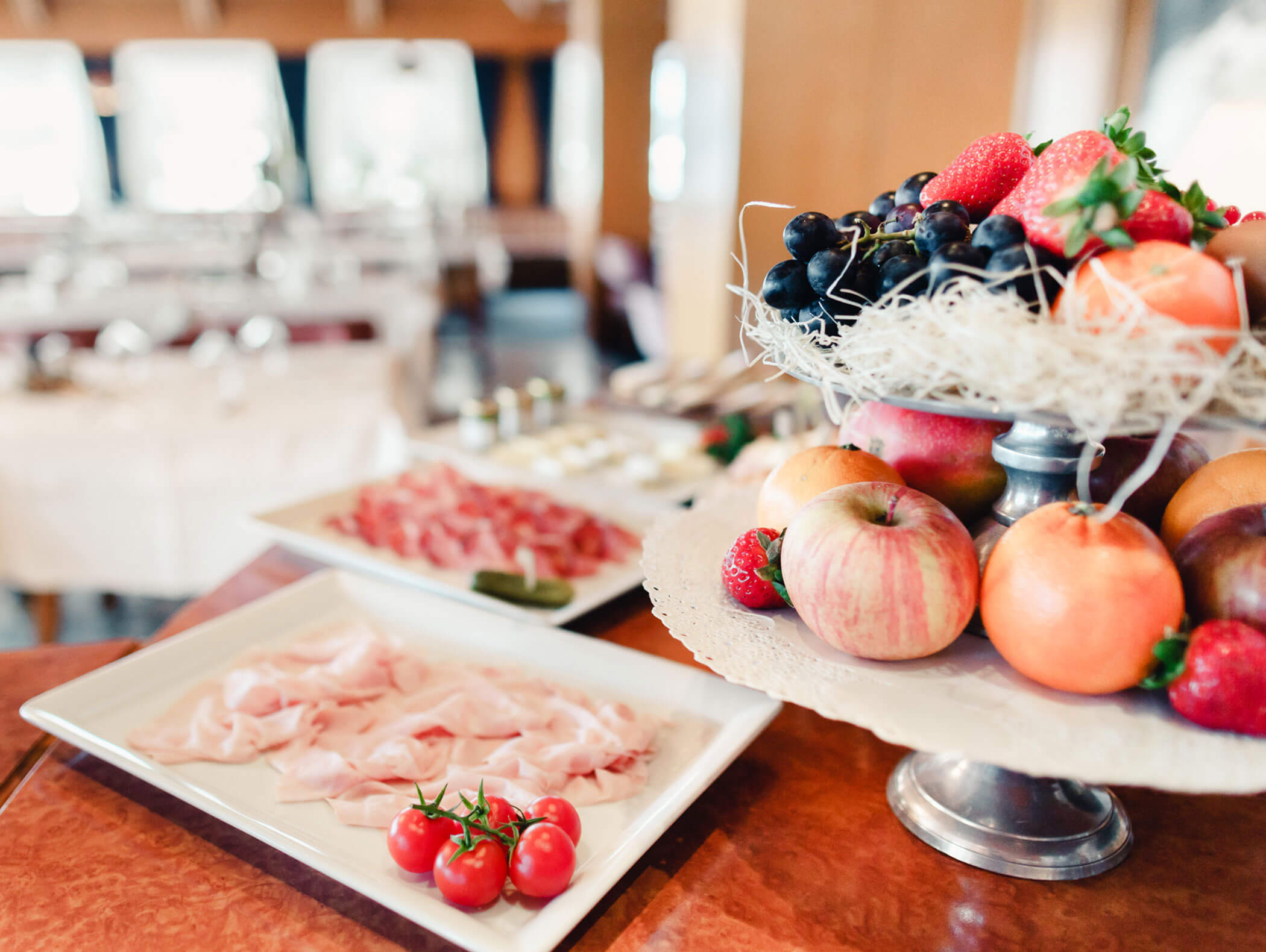 A fruit stand with apples and berries beside plates of sliced meats and cherry tomatoes in a bright dining room. - Hotel Kristiania