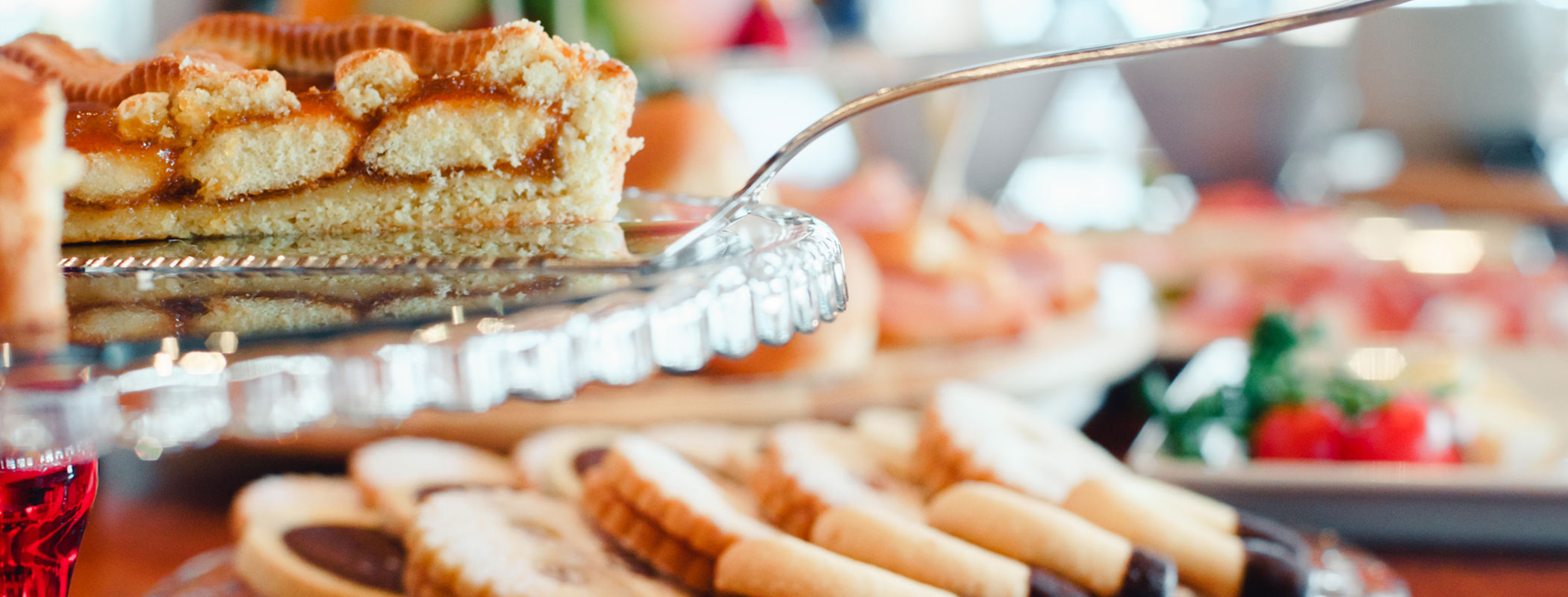 A close-up of a pie slice being served, with cookies and pastries visible on a table in the background. - Hotel Kristiania