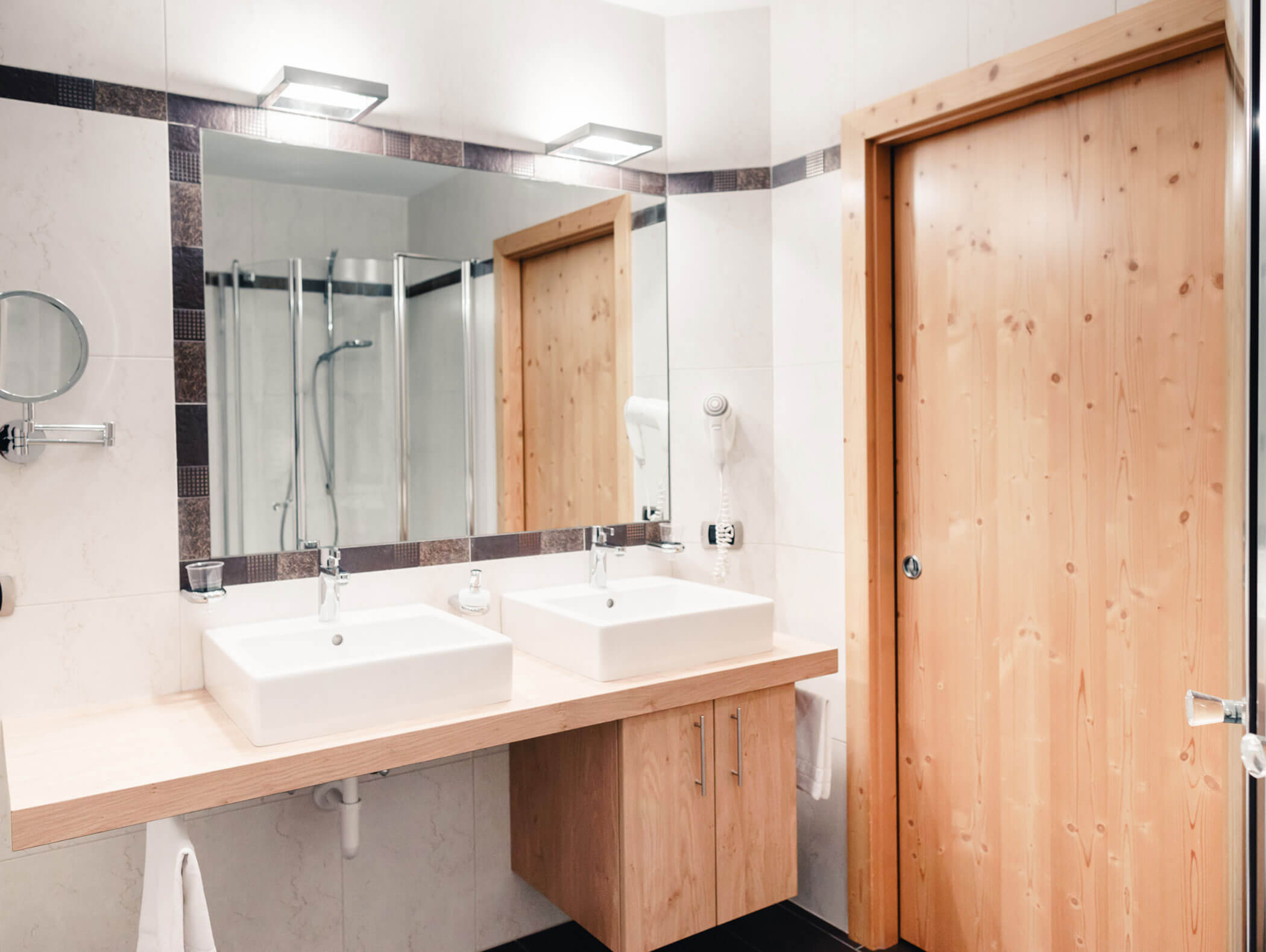 Bagno moderno con doppio lavabo, ampio specchio, porta in legno e box doccia in vetro sullo sfondo. - Hotel Kristiania