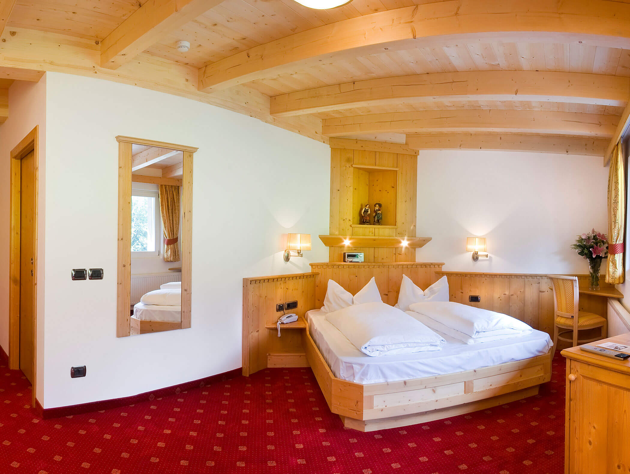 Accogliente camera d'albergo con letto matrimoniale in legno, moquette rossa, specchio a parete e luce naturale che entra dalla finestra. - Hotel Kristiania