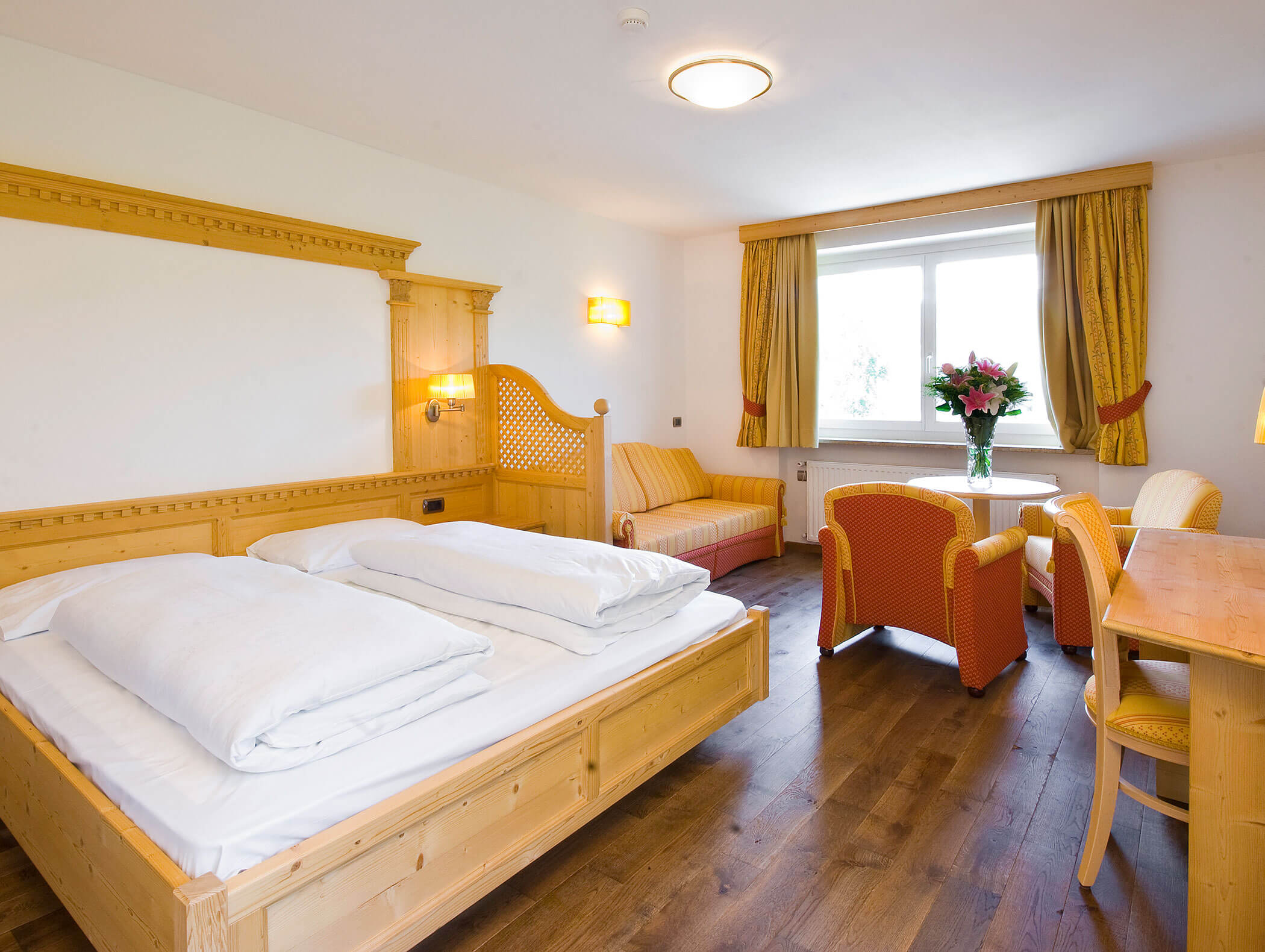 Una luminosa camera d'albergo con un letto in legno, sedie arancioni, un divano e una finestra con tende gialle e fiori. - Hotel Kristiania