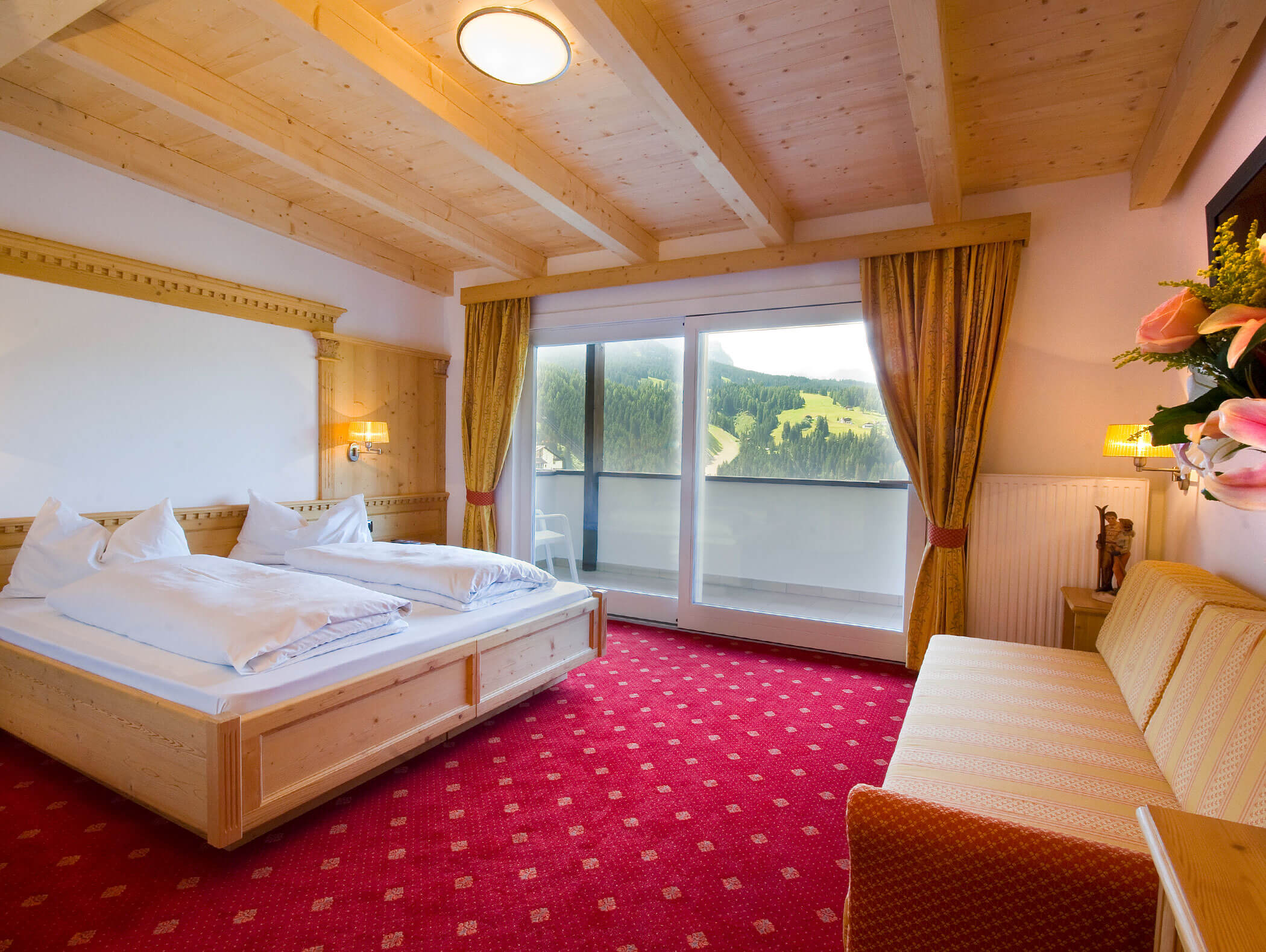 Accogliente camera d'albergo con letto matrimoniale, divano, moquette rossa, soffitto in legno e balcone con vista sulle verdi colline. - Hotel Kristiania