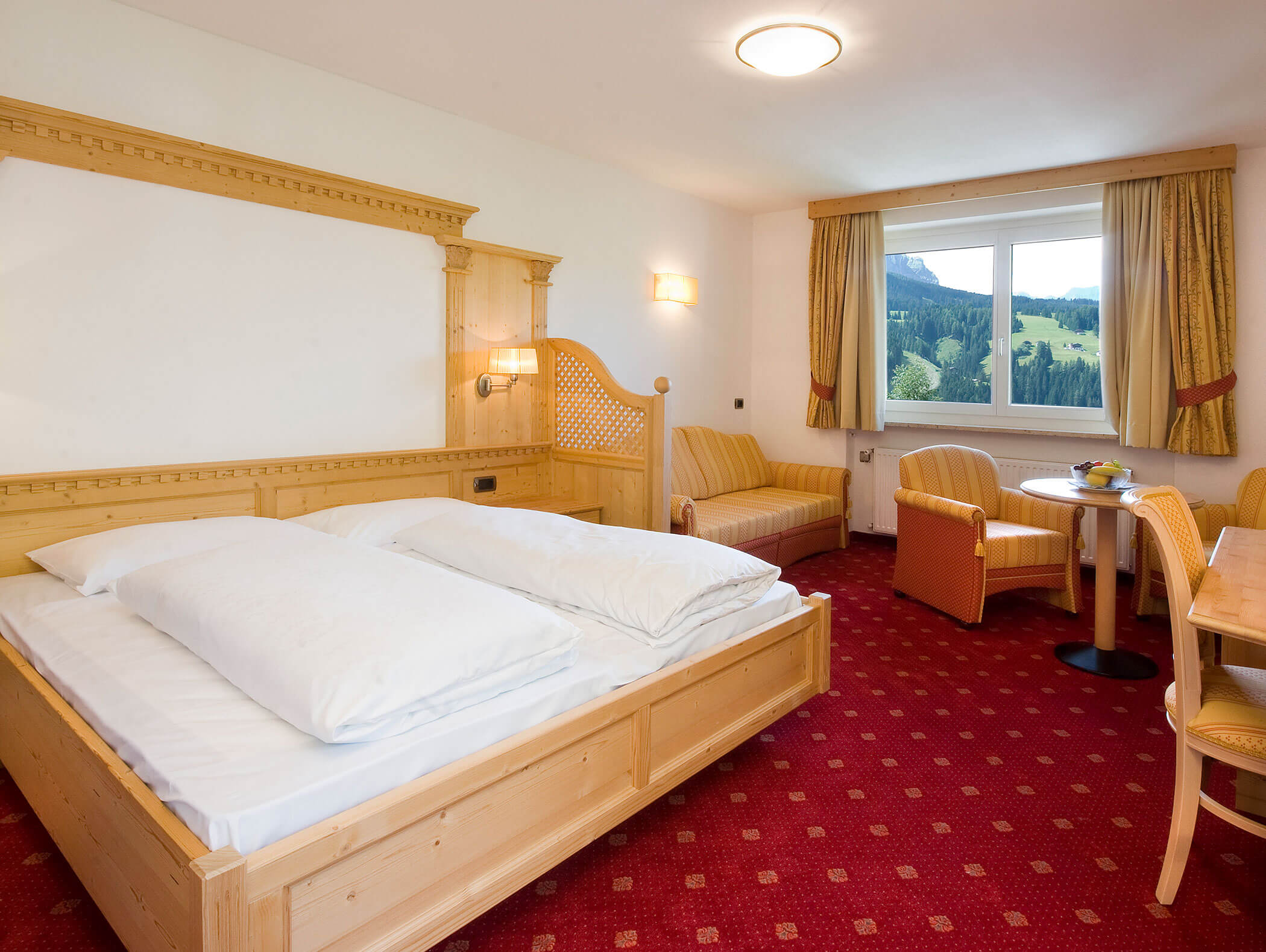 Camera d'albergo con letto in legno, moquette rossa, salottino, tavolo e finestra con vista panoramica. - Hotel Kristiania