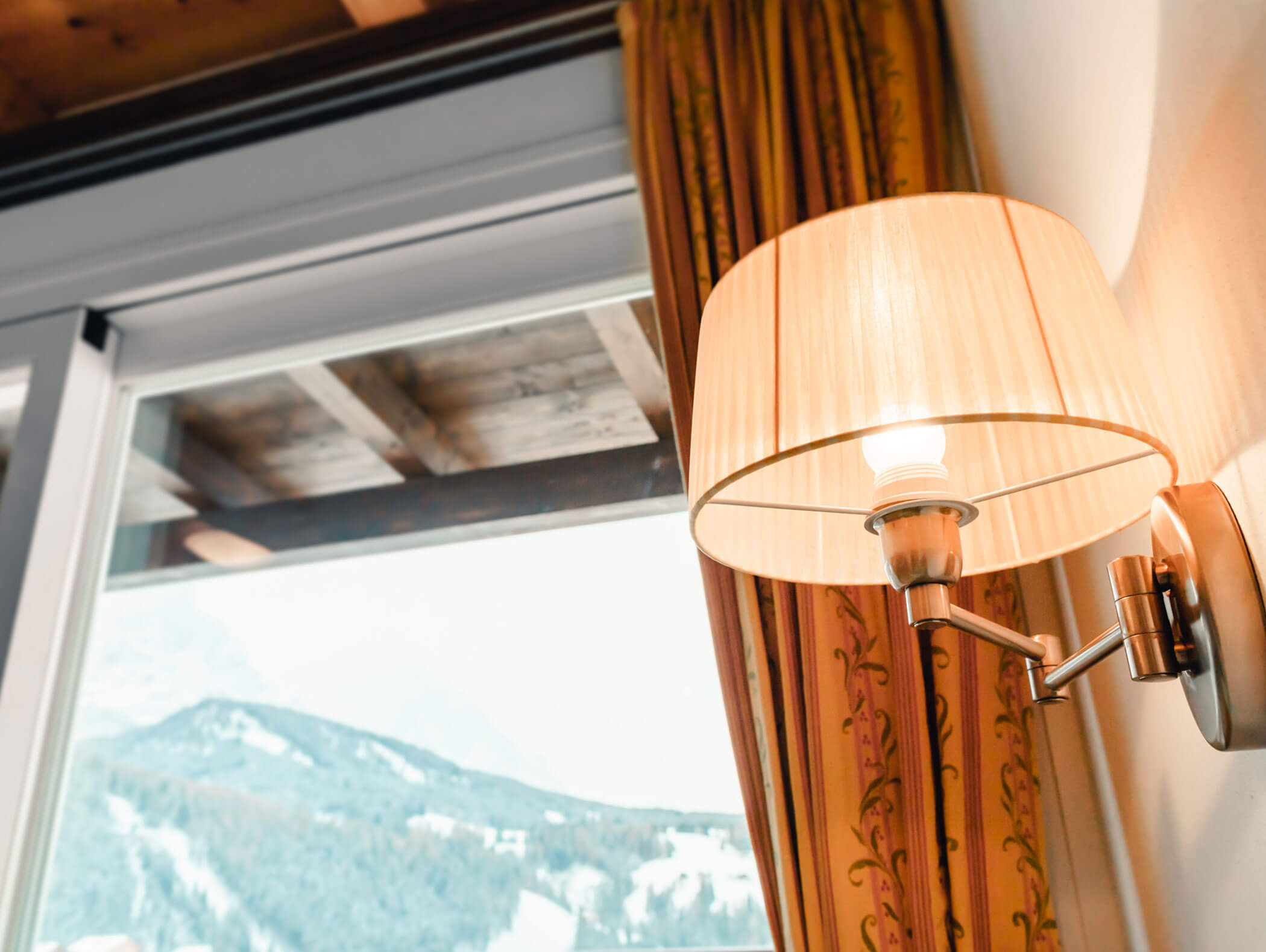 Lampada da parete con una lampadina accesa accanto a una finestra affacciata su montagne innevate e una tenda a motivi decorativi. - Hotel Kristiania