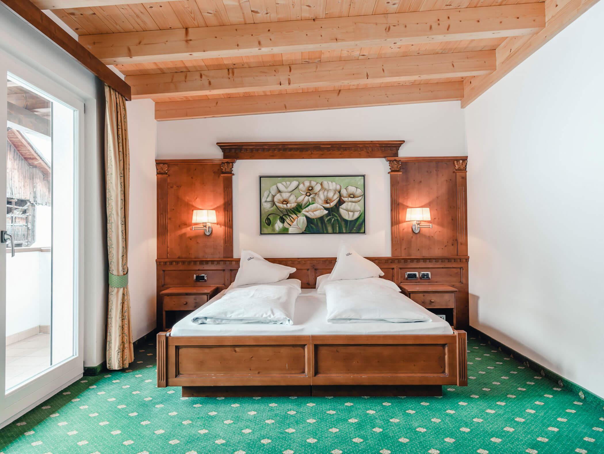 Accogliente camera da letto con un letto in legno, un tappeto verde, un quadro con motivi floreali e due applique sotto un soffitto in legno. - Hotel Kristiania