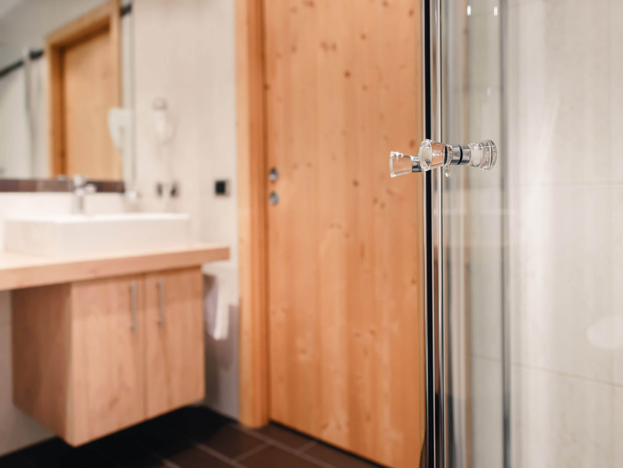 Primo piano della maniglia di una porta doccia in vetro in un bagno moderno con ante in legno e un lavabo. - Hotel Kristiania
