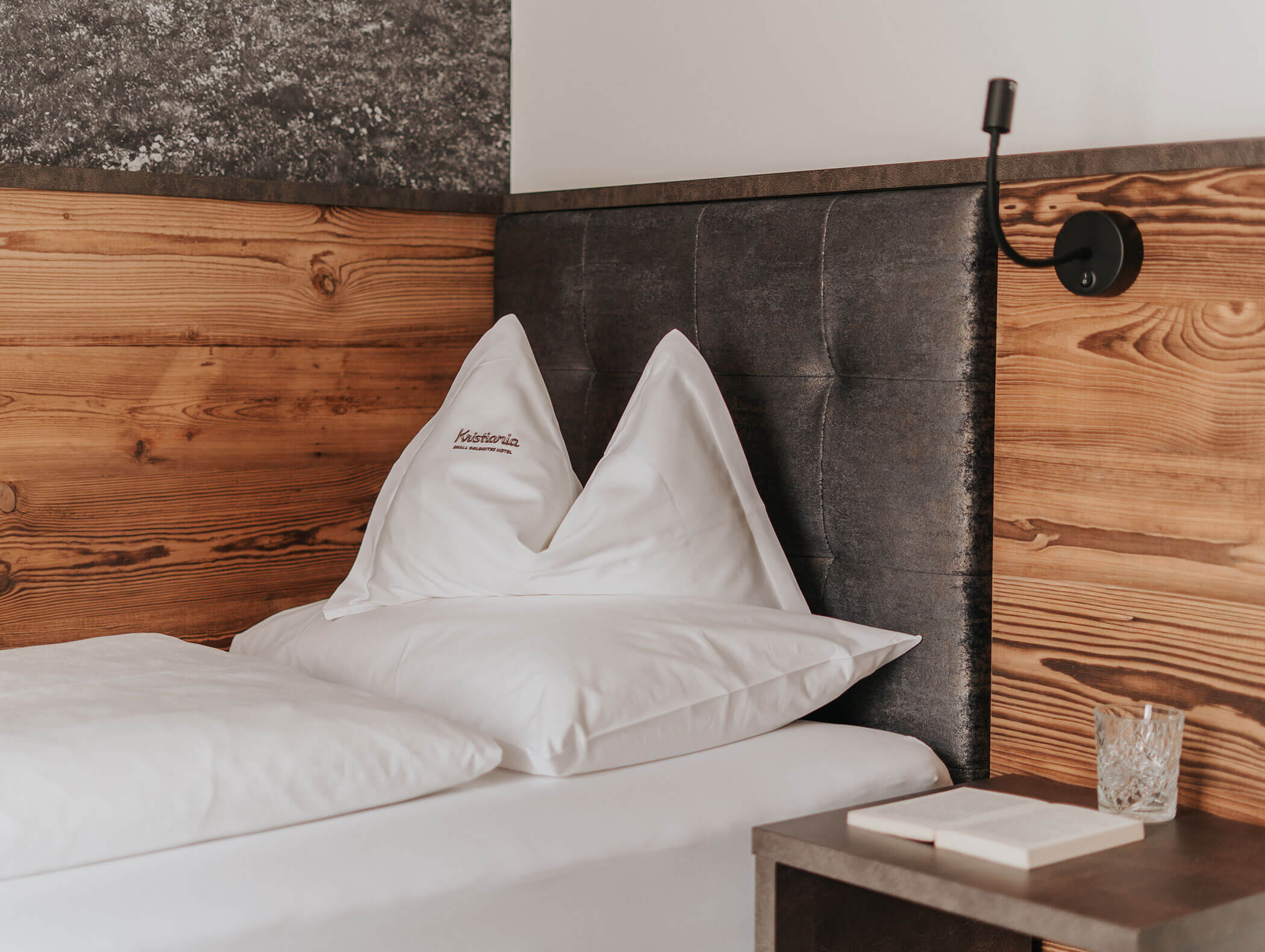 Un letto ben rifatto con lenzuola bianche e cuscini, accanto a un comodino di legno su cui sono appoggiati un bicchiere e un libro aperto. - Hotel Kristiania