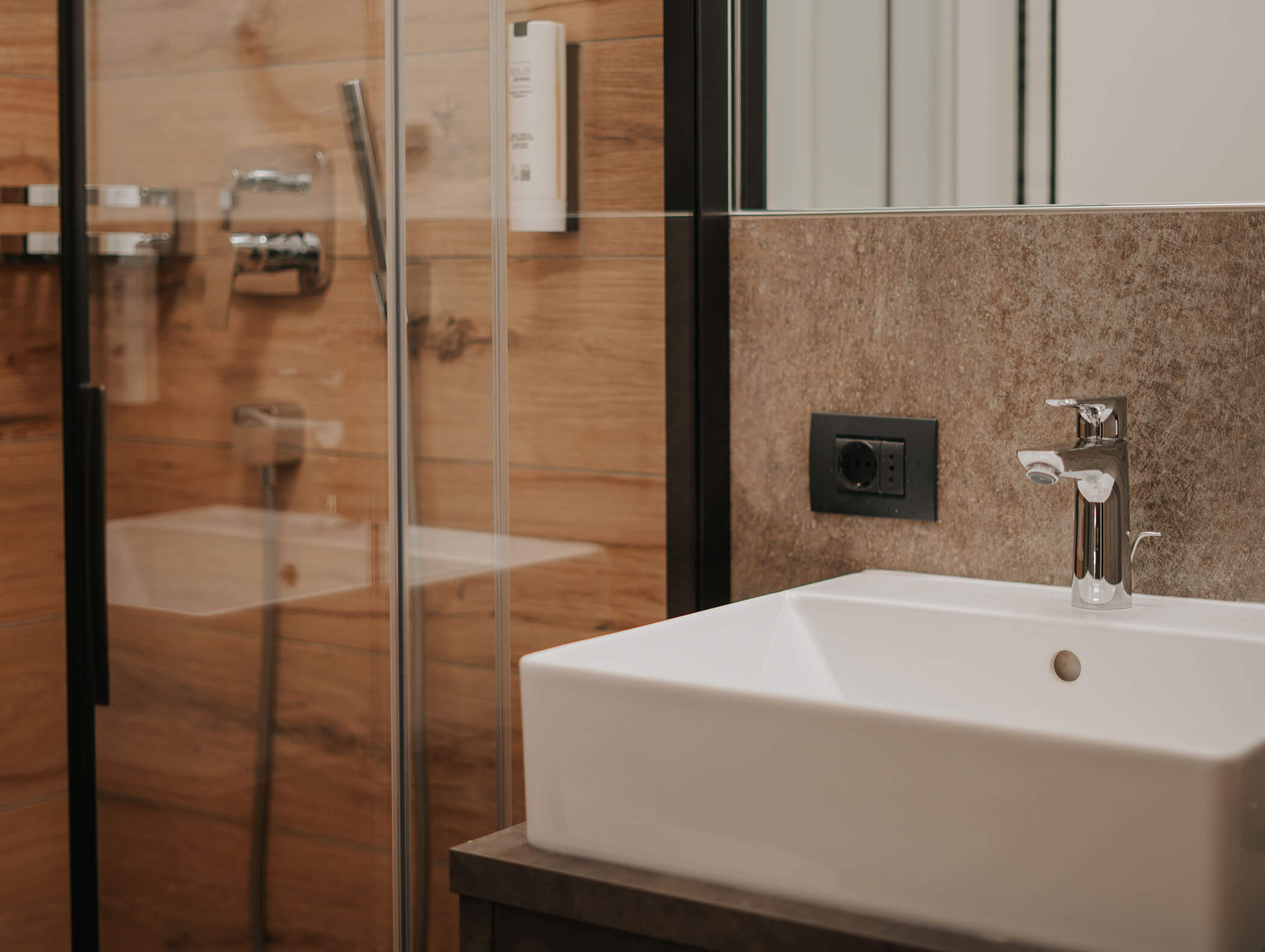 Bagno moderno con lavabo bianco, rubinetto cromato, box doccia in vetro e pareti con inserti in legno. - Hotel Kristiania