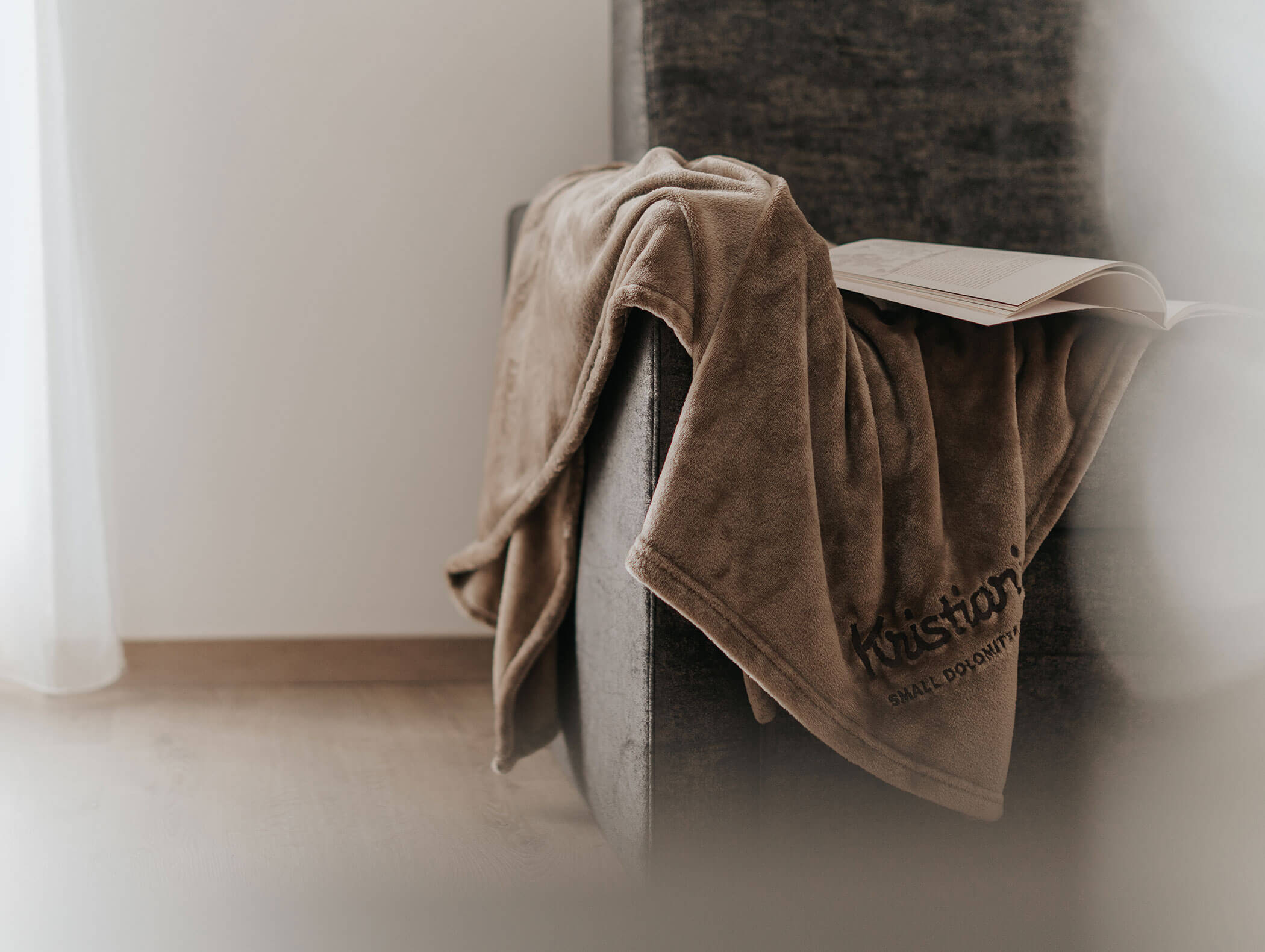 Una morbida coperta marrone e un libro aperto appoggiato sul bracciolo di una poltrona grigia in una stanza accogliente e inondata di luce. - Hotel Kristiania