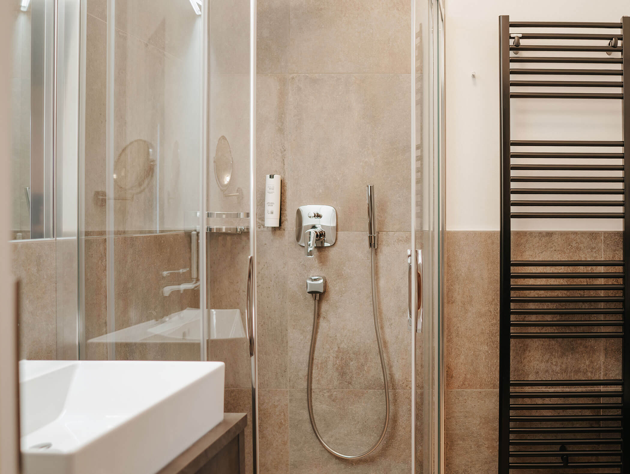 Doccia moderna con porte in vetro, rubinetteria a parete e un scaldasalviette nero in un bagno piastrellato di beige. - Hotel Kristiania