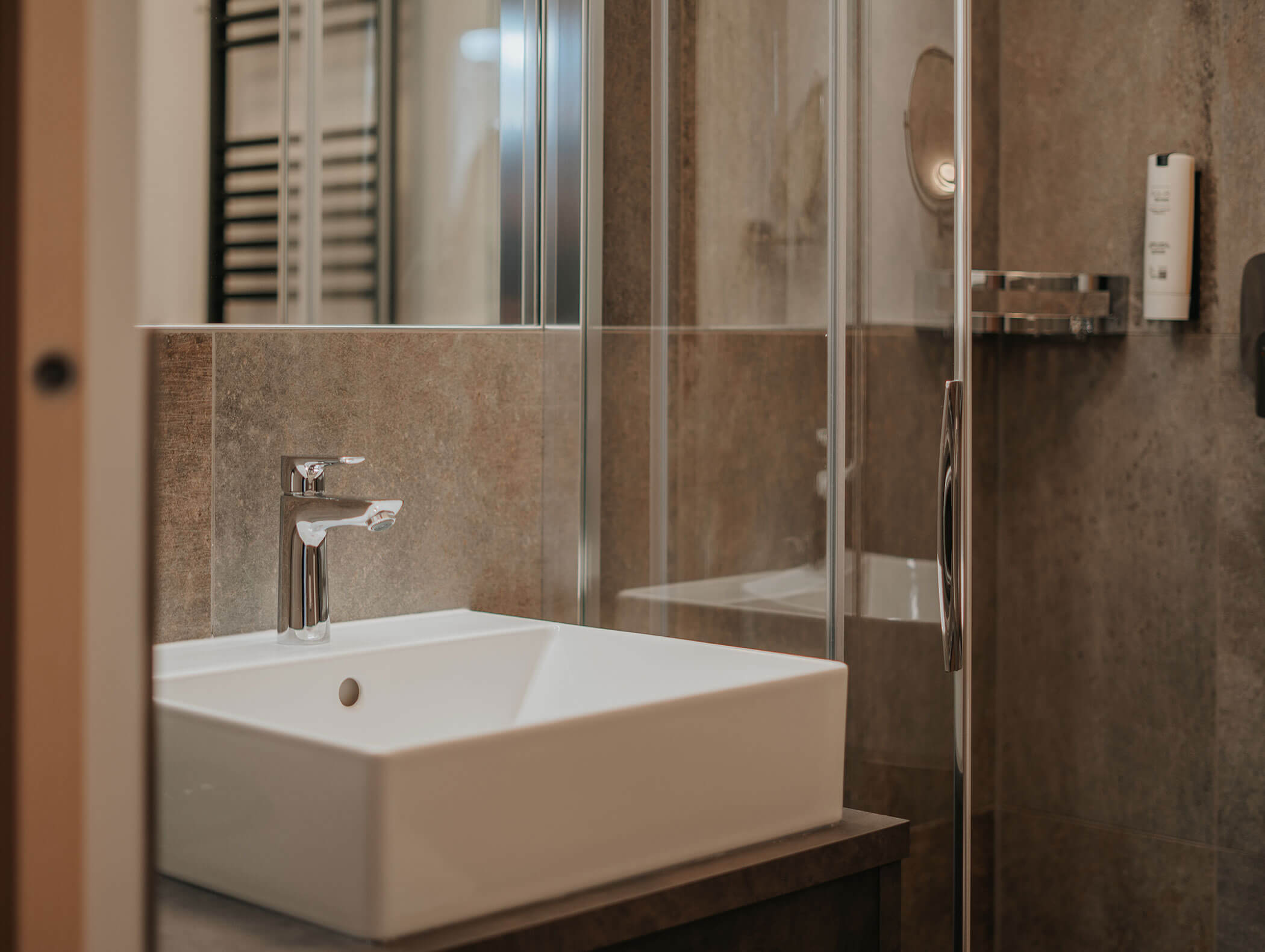 Bagno moderno con lavabo quadrato bianco, rubinetto cromato e box doccia in vetro su pareti rivestite di piastrelle marroni. - Hotel Kristiania