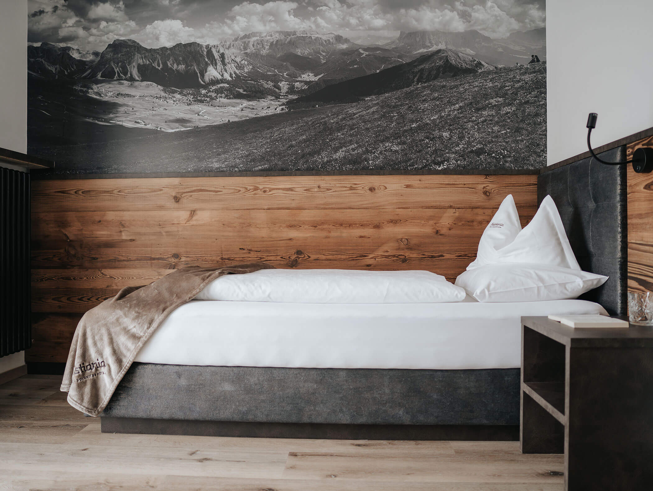 Un letto singolo ben rifatto, con lenzuola bianche e un copriletto di pelliccia, addossato a una parete rivestita di pannelli di legno e decorata con un murale che raffigura una montagna. - Hotel Kristiania