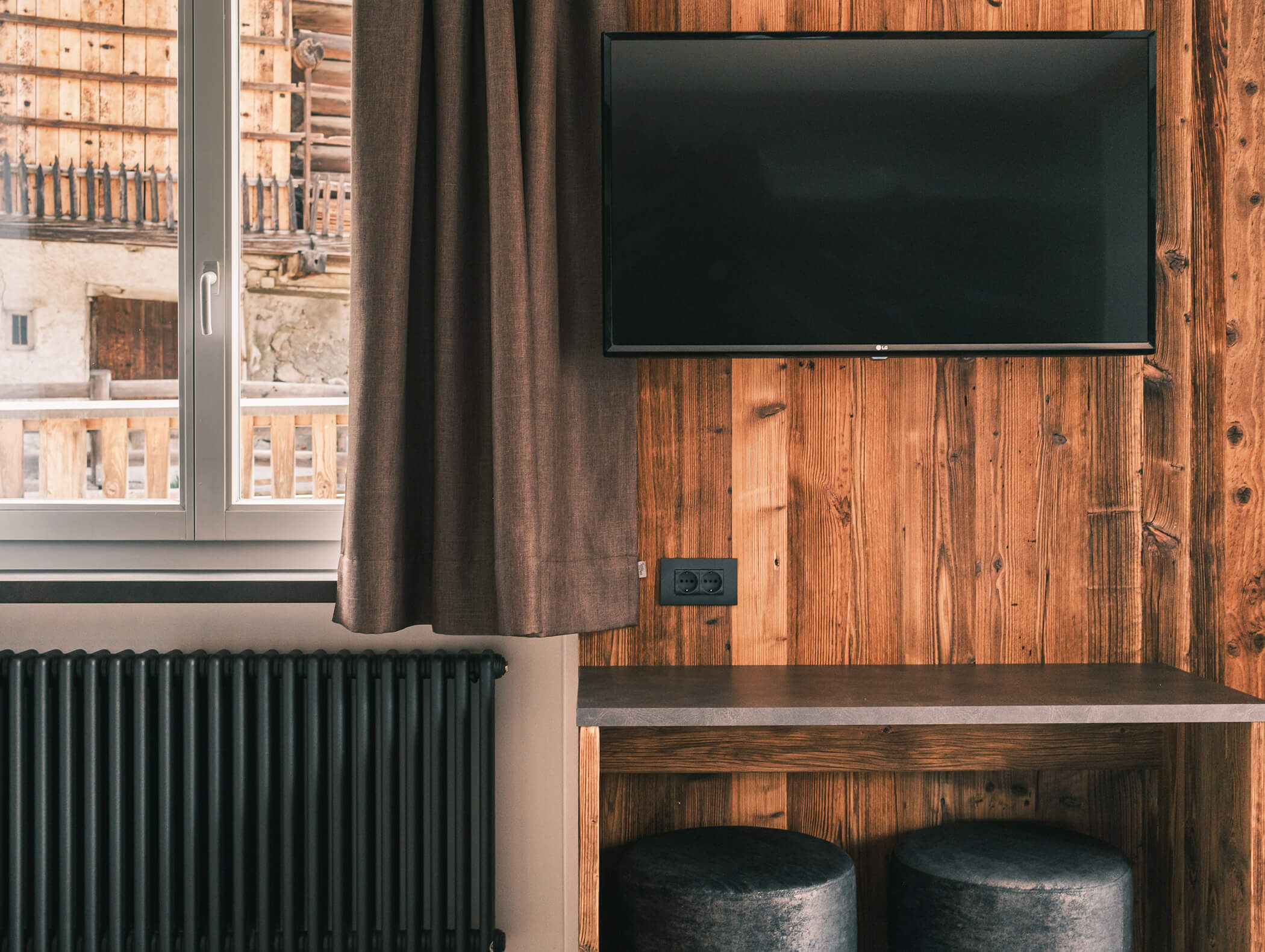 Una TV appesa alla parete sopra una scrivania di legno con due sgabelli accanto a una finestra coperta da una tenda marrone. - Hotel Kristiania