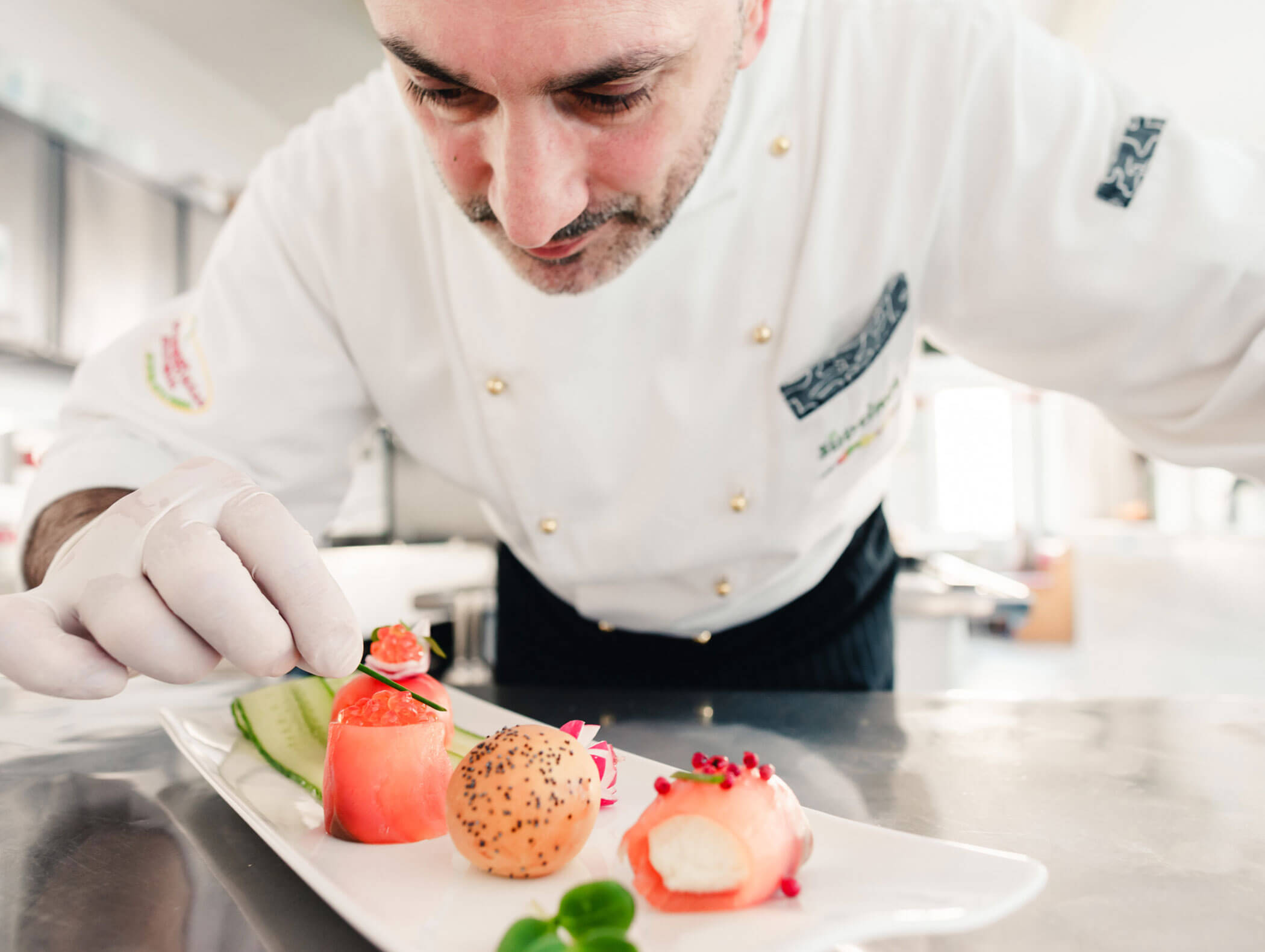 Ein Koch in weißer Uniform richtet in einer hellen Küche sorgfältig Gourmet-Häppchen auf einem weißen Teller an. - Hotel Kristiania