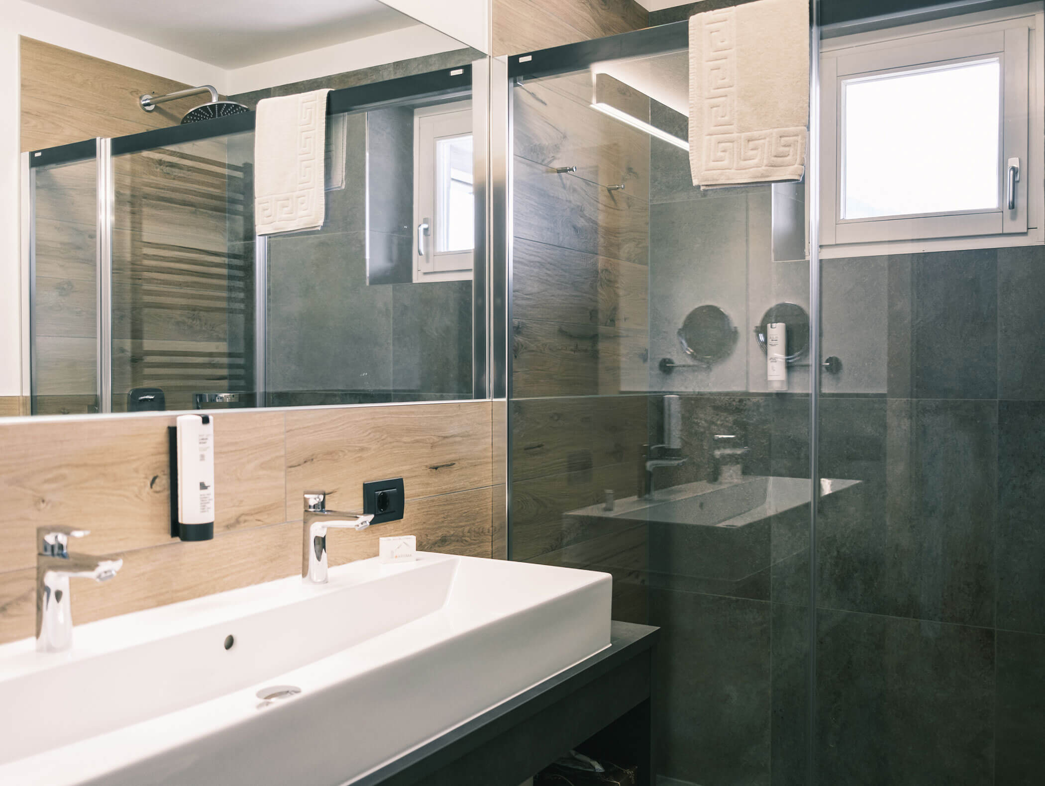 Modernes Badezimmer mit Doppelwaschbecken, großem Spiegel, Glasduschkabine und Wänden in Holzoptik; darüber hängt ein Handtuch. - Hotel Kristiania