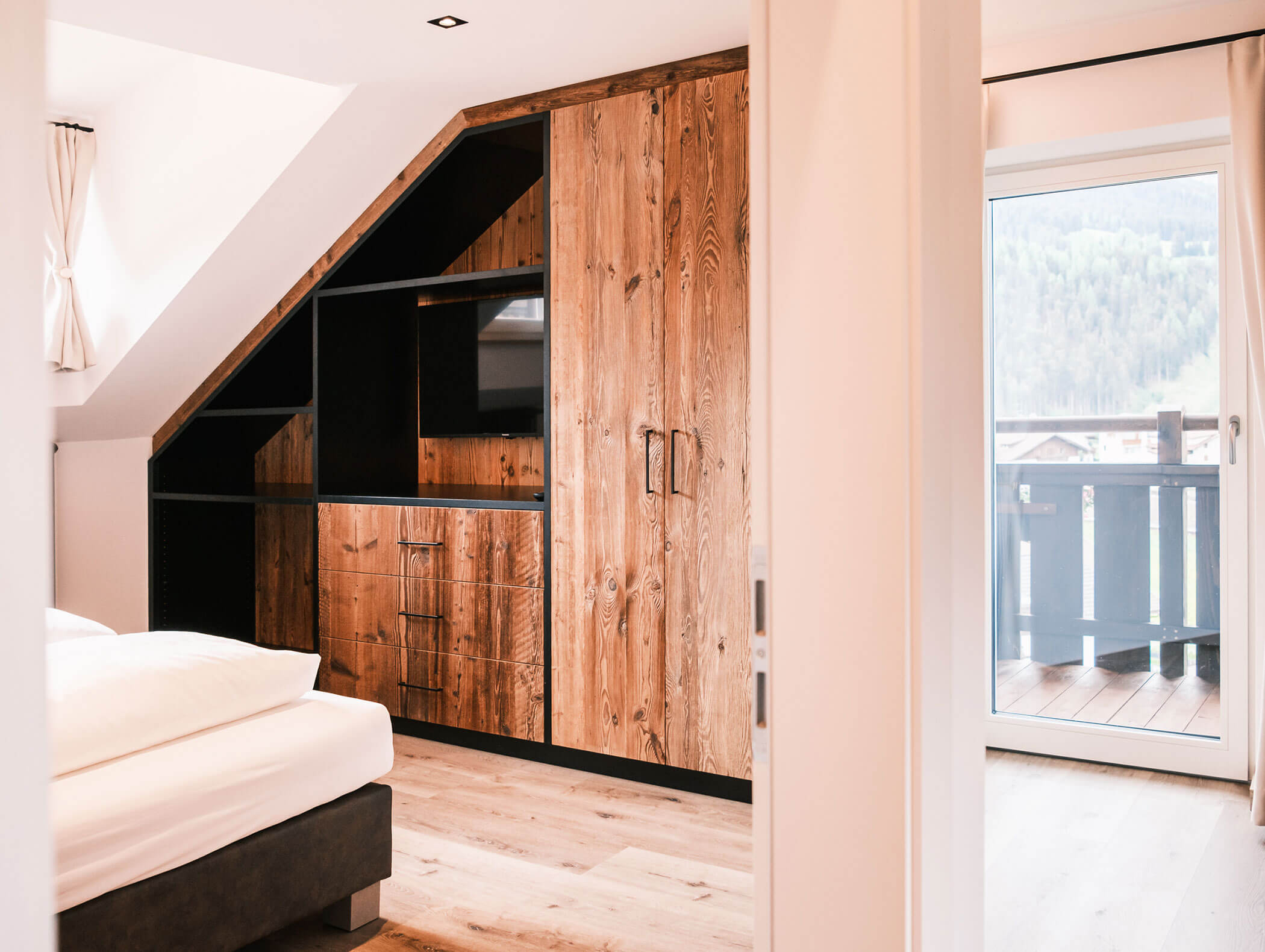 Camera da letto moderna con soffitto spiovente, armadio a muro in legno, letto e una porta che conduce a un balcone con vista. - Hotel Kristiania