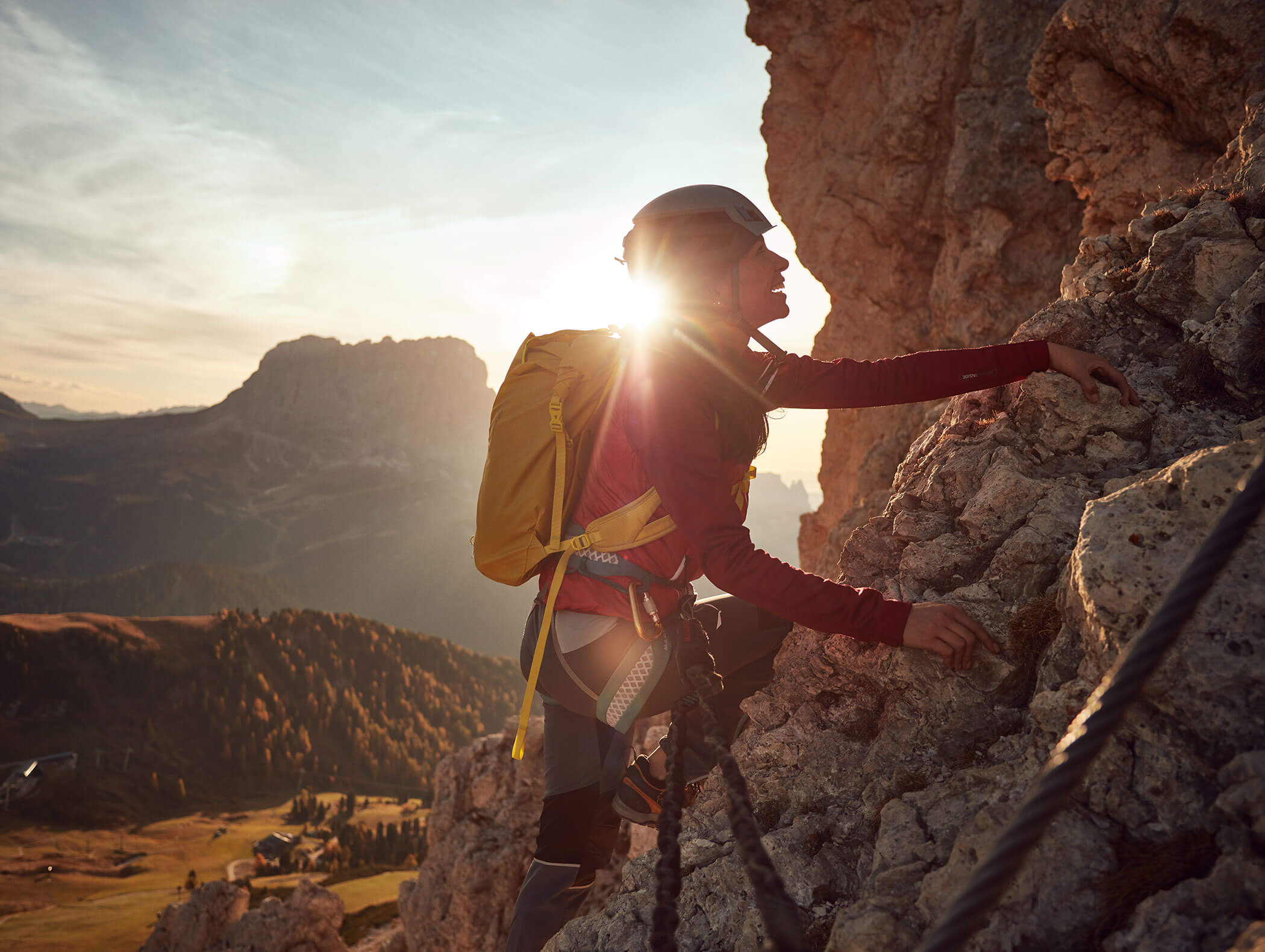 Ein Kletterer mit einem gelben Rucksack erklimmt bei Sonnenuntergang eine felsige Klippe, im Hintergrund sind Berge und ein Tal zu sehen. - Hotel Kristiania