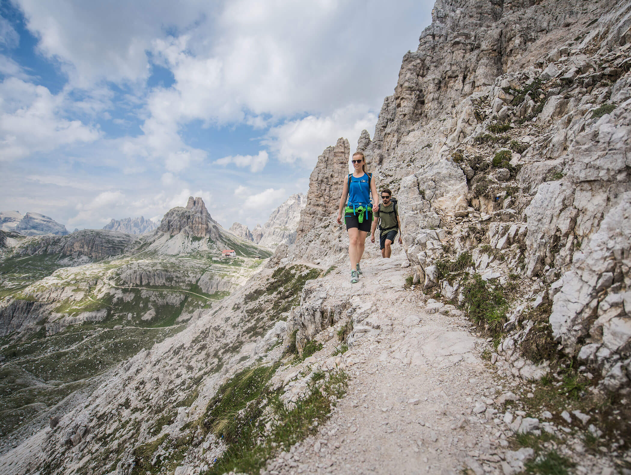 Zwei Menschen wandern auf einem felsigen Bergpfad, im Hintergrund sind zerklüftete Gipfel und ein teilweise bewölkter Himmel zu sehen. - Hotel Kristiania