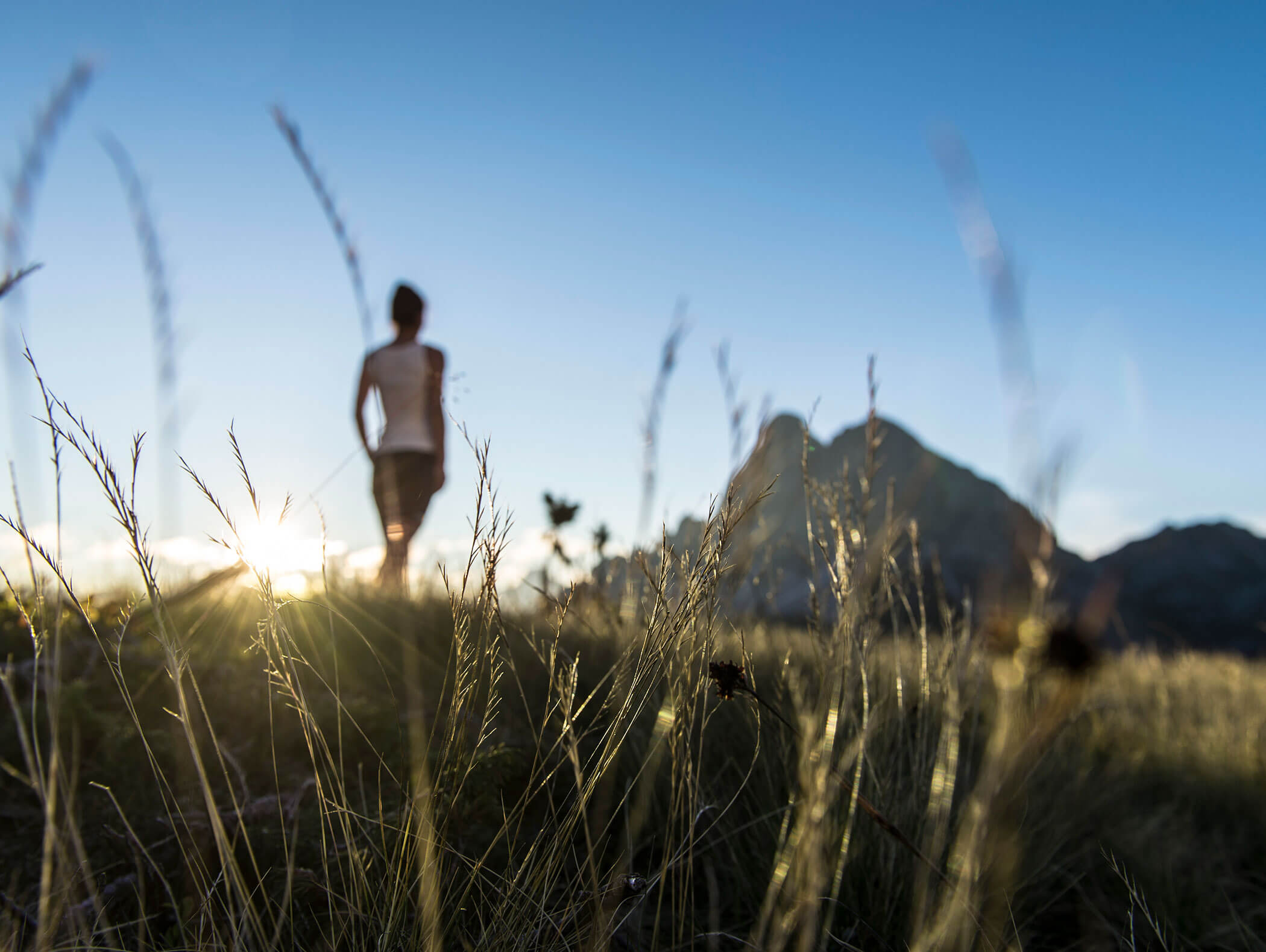 Eine Person steht bei Sonnenuntergang auf einem grasbewachsenen Hügel, im Hintergrund sind Berge zu sehen, das Gras im Vordergrund ist unscharf. - Hotel Kristiania