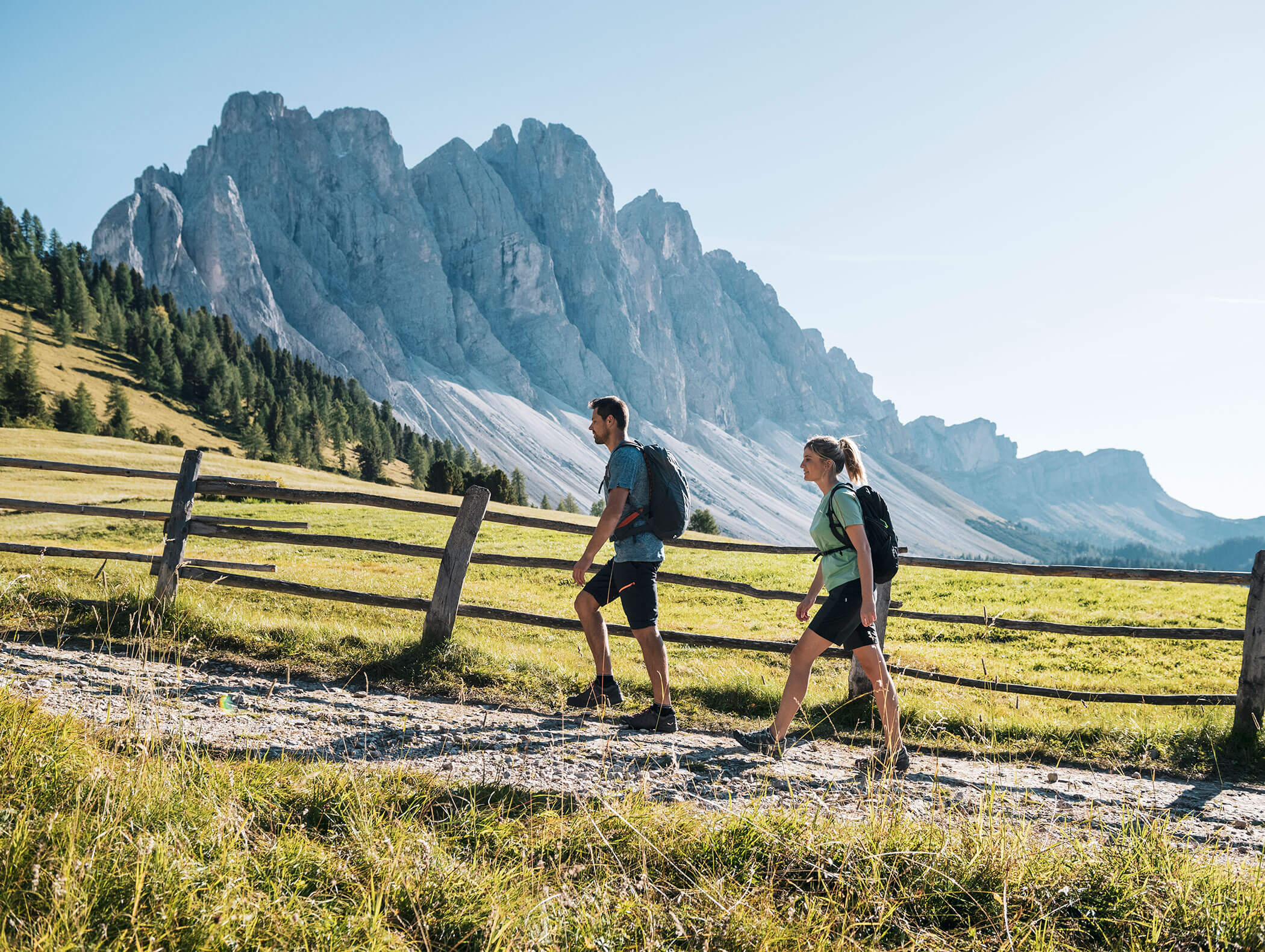 Zwei Menschen wandern auf einem Feldweg neben einem Holzzaun, im Hintergrund sind Berge zu sehen, über ihnen erstreckt sich ein klarer Himmel. - Hotel Kristiania
