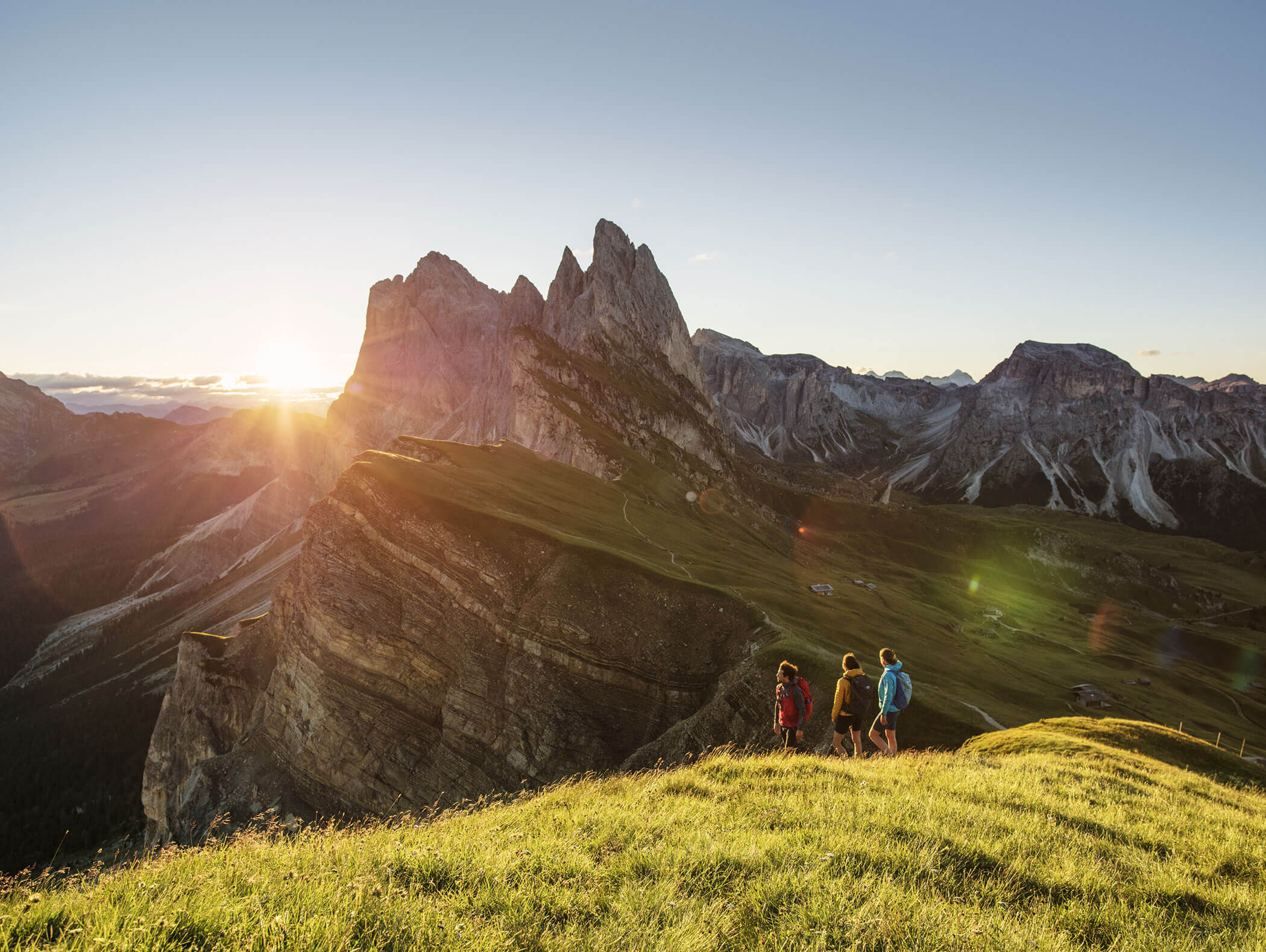 Drei Wanderer spazieren bei Sonnenaufgang auf einem grasbewachsenen Hügel und blicken auf beeindruckende Berggipfel und Täler. - Hotel Kristiania