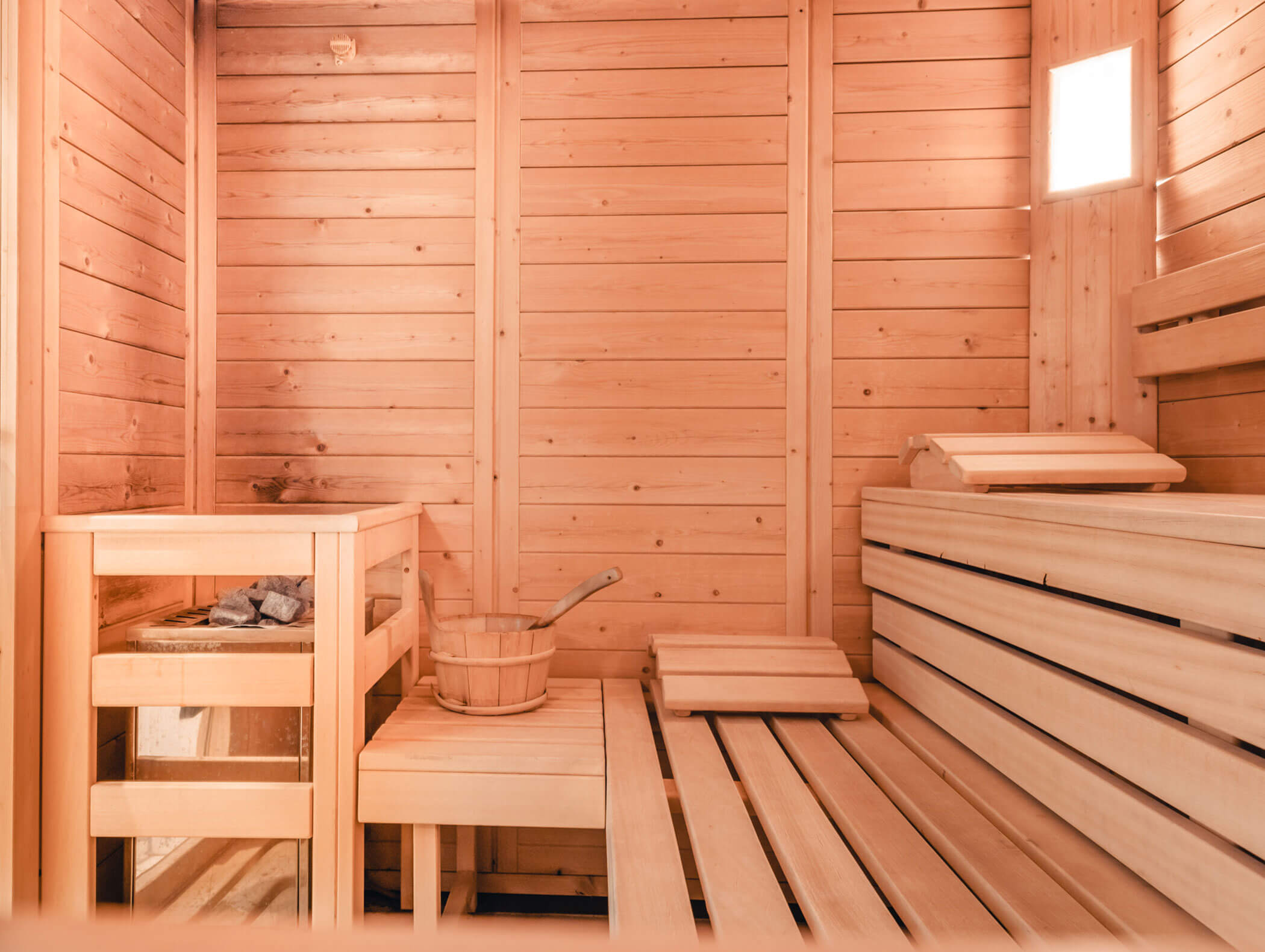 Innenansicht einer Holzsauna mit Bänken, einem Eimer, einer Schöpfkelle und heißen Steinen bei warmer Beleuchtung. - Hotel Kristiania
