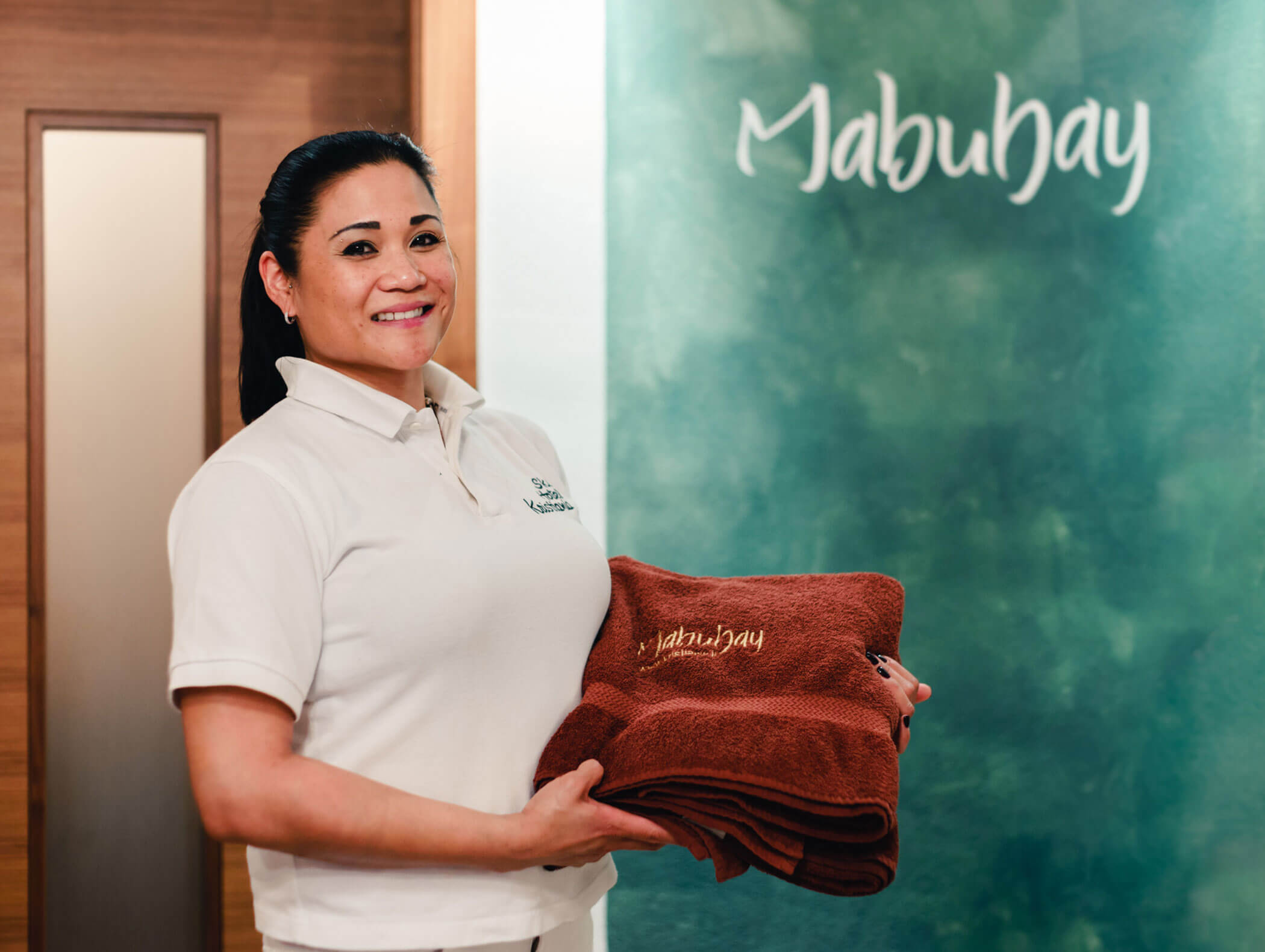 Lächelnde Frau in Uniform, die gefaltete braune Handtücher vor einer grünen Wand hält, auf der „Mabuhay“ steht. - Hotel Kristiania
